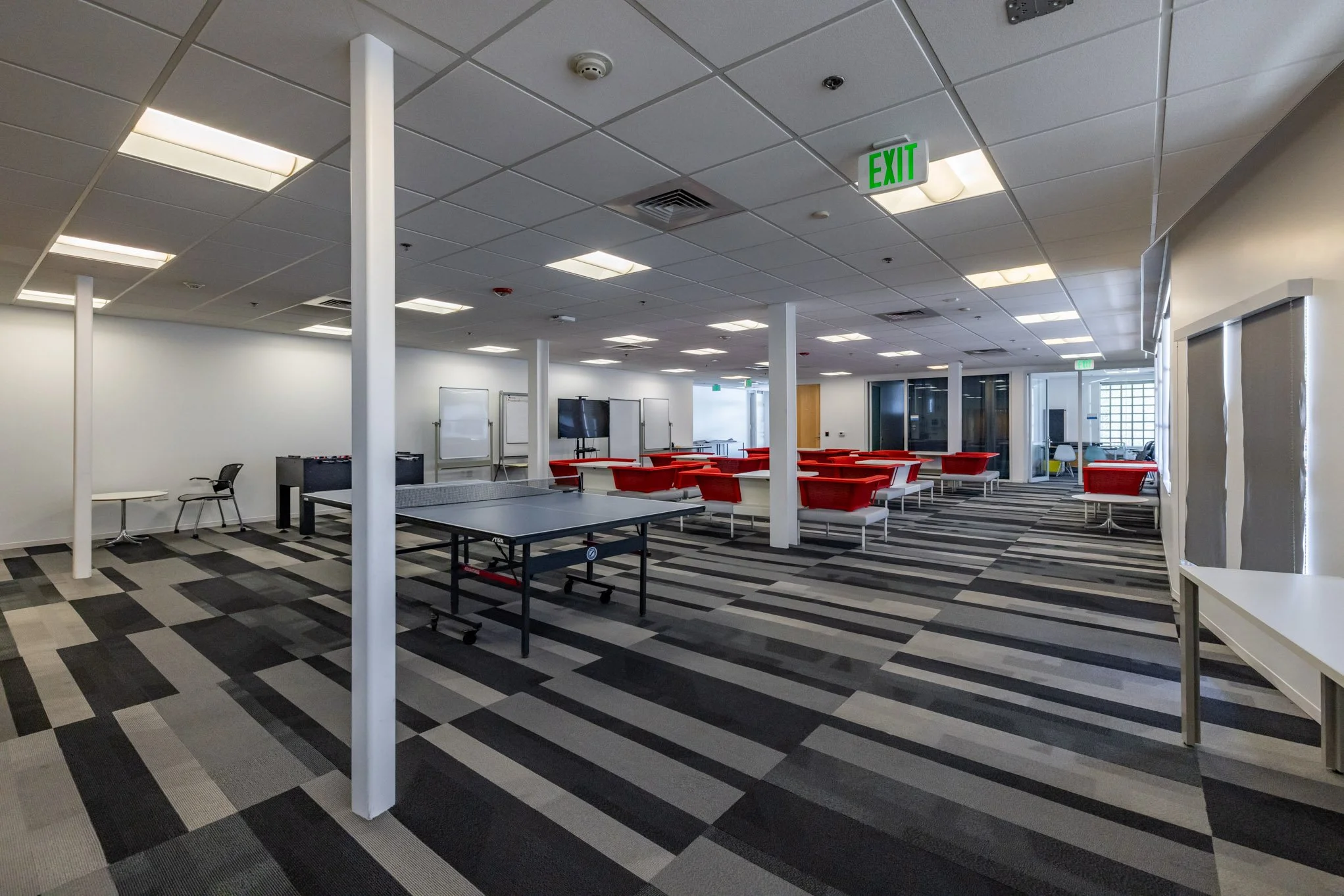 Empty modern office lounge area with red chairs, white tables, a ping pong table, whiteboards, and a large flat-screen TV on a patterned carpet floor.
