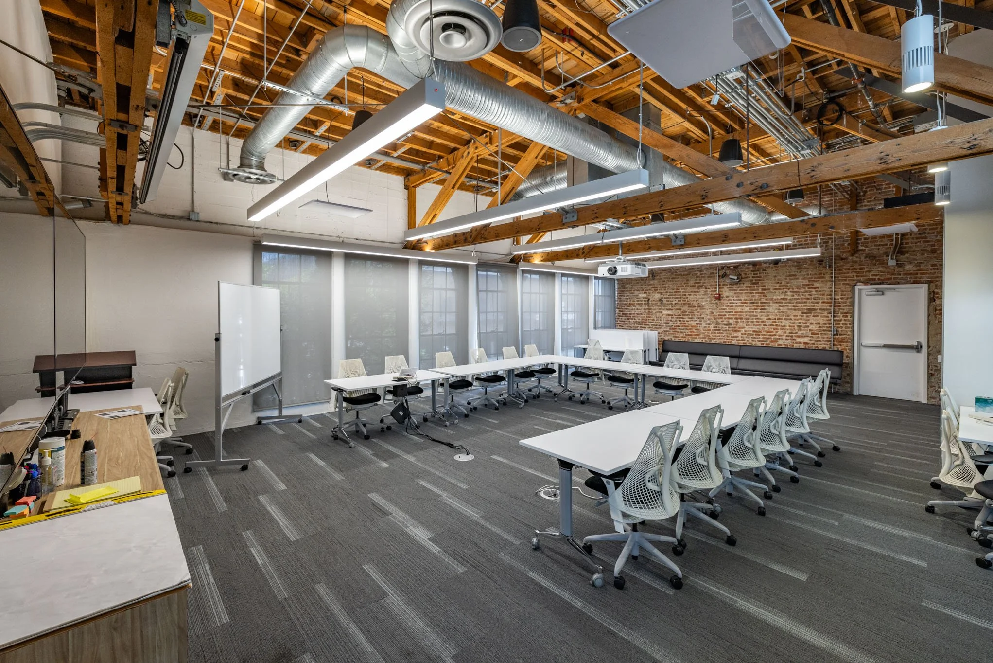 Modern conference room with white tables arranged in a U-shape, white mesh chairs, a whiteboard, a projector, and exposed wooden beams and brick walls.