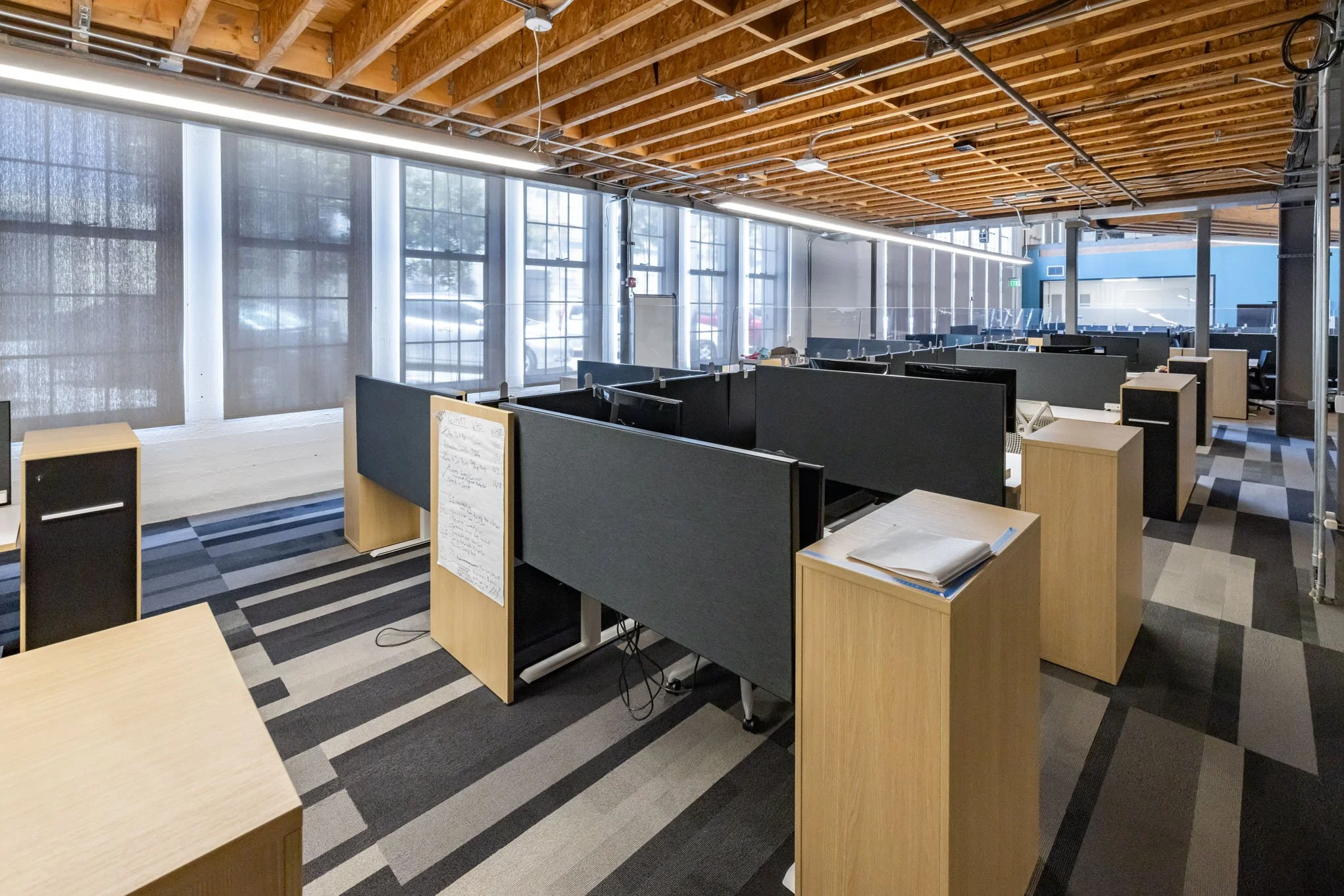 Empty modern office space with wooden desks, black dividers, and large windows with blinds, exposed wooden ceiling beams, and patterned carpet.