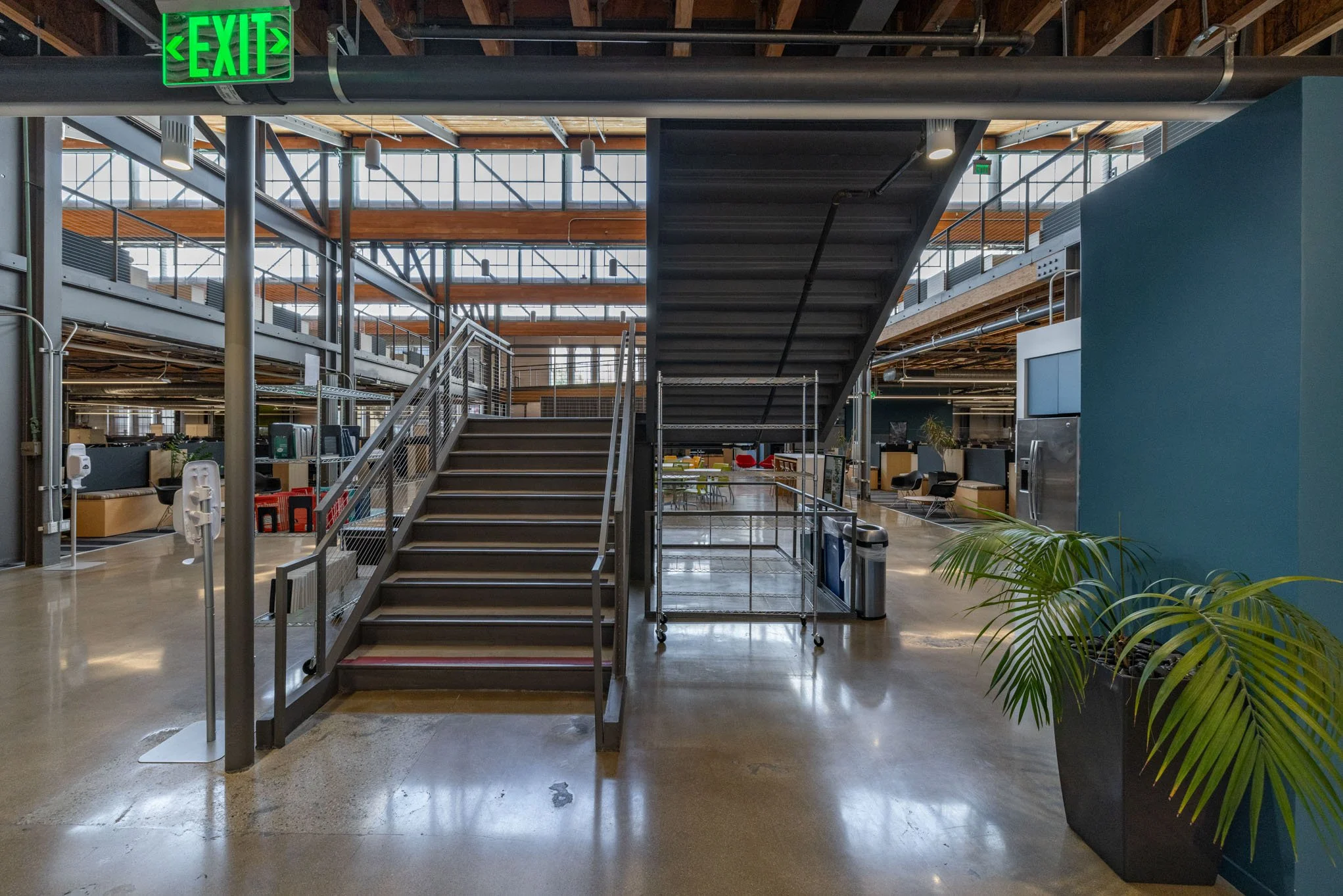 Interior of a modern office space with a staircase, green exit sign, and various work areas in the background, with polished concrete floors and large windows.