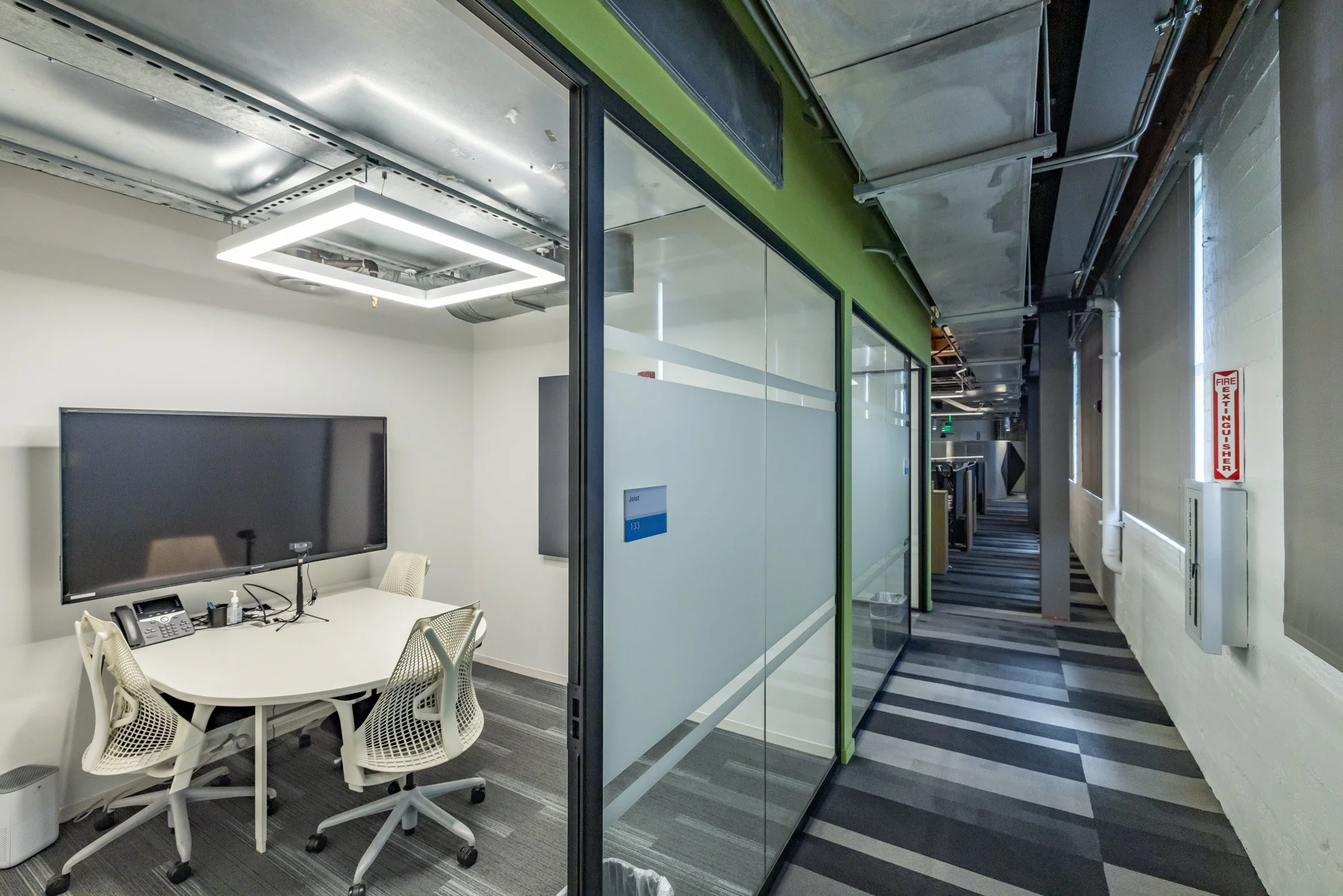 Modern office conference room with white table, four white chairs, large wall-mounted screen, glass walls, and a corridor with striped carpet and fire extinguisher sign.