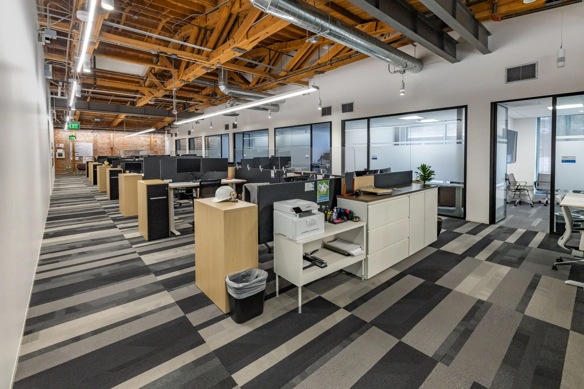 An open-plan office space with multiple desks, partition dividers, and chairs, featuring exposed wooden ceiling beams and ductwork, glass-walled meeting rooms, and a patterned carpet.