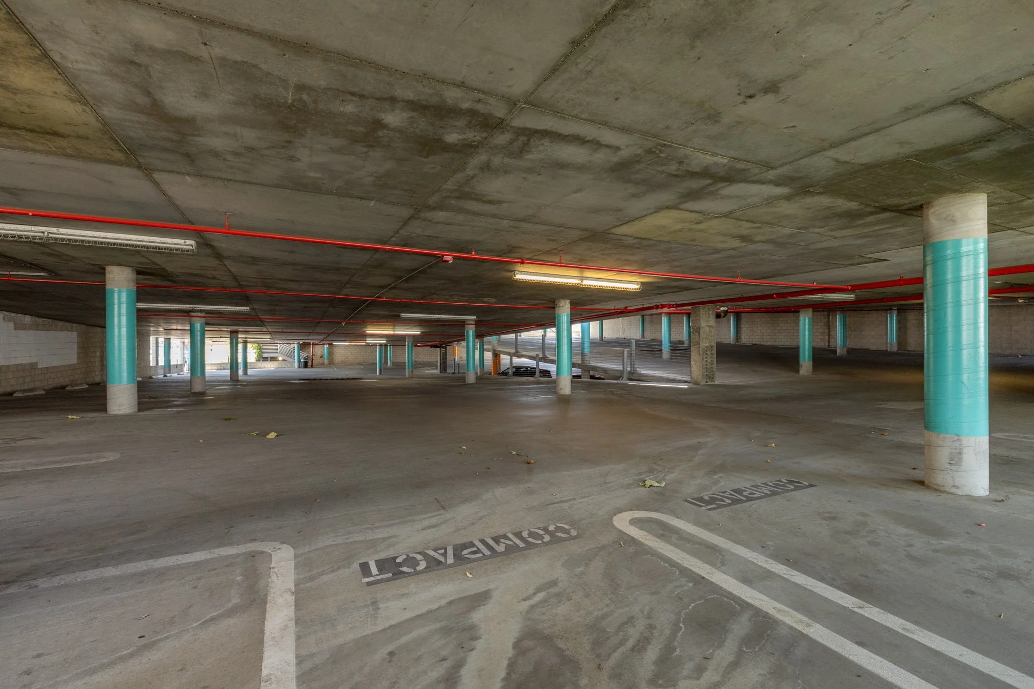 Underground parking garage with concrete pillars wrapped in teal and red pipes along the ceiling, empty parking spots marked with 'RESERVED' signs, and natural light coming from openings in the walls.