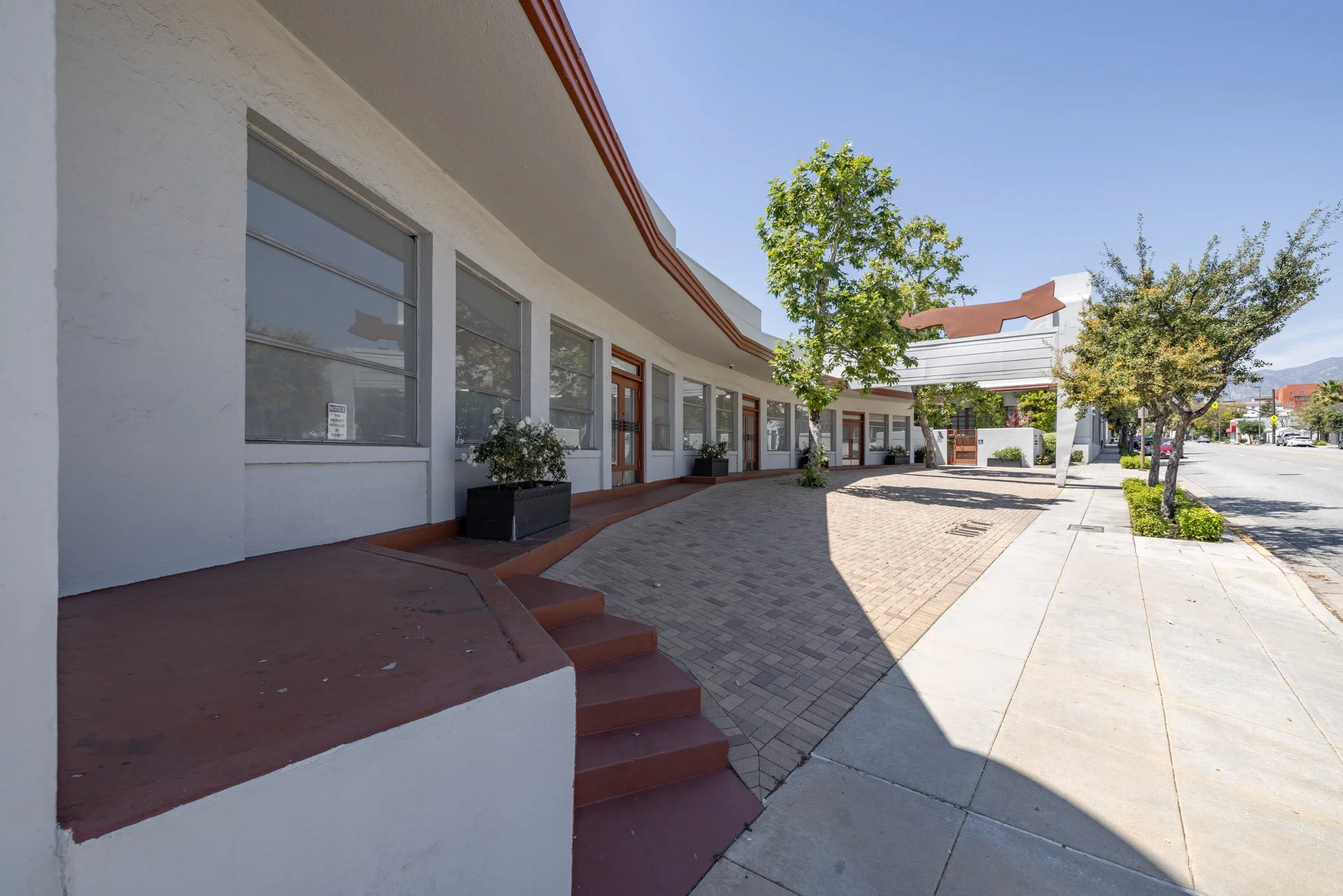 Empty sidewalk and storefront with trees, planters, and a curved building facade under a clear blue sky.