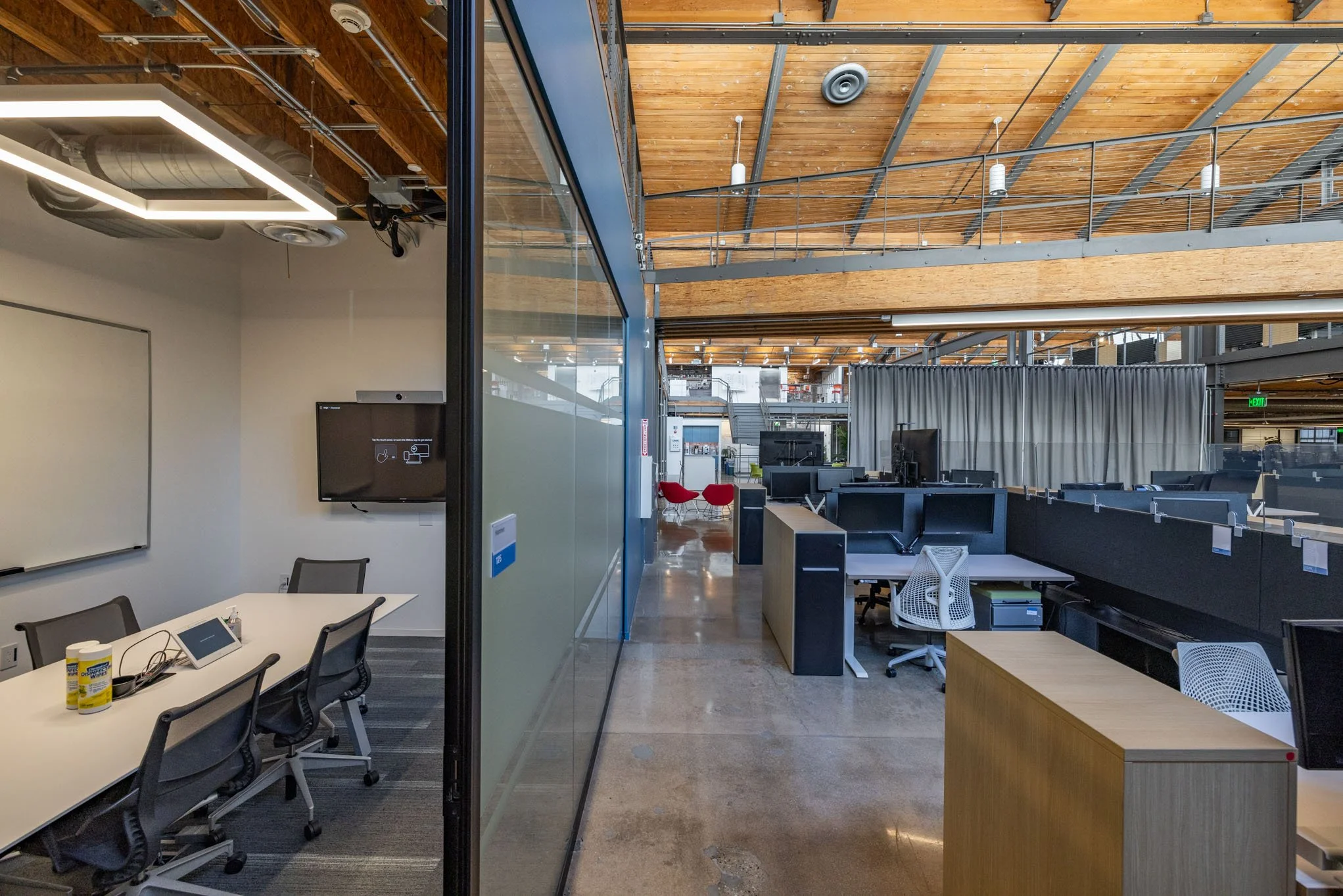 Interior of a modern office space with open work areas, desks, and chairs, with a glass wall dividing the space and a wooden ceiling with metal beams.