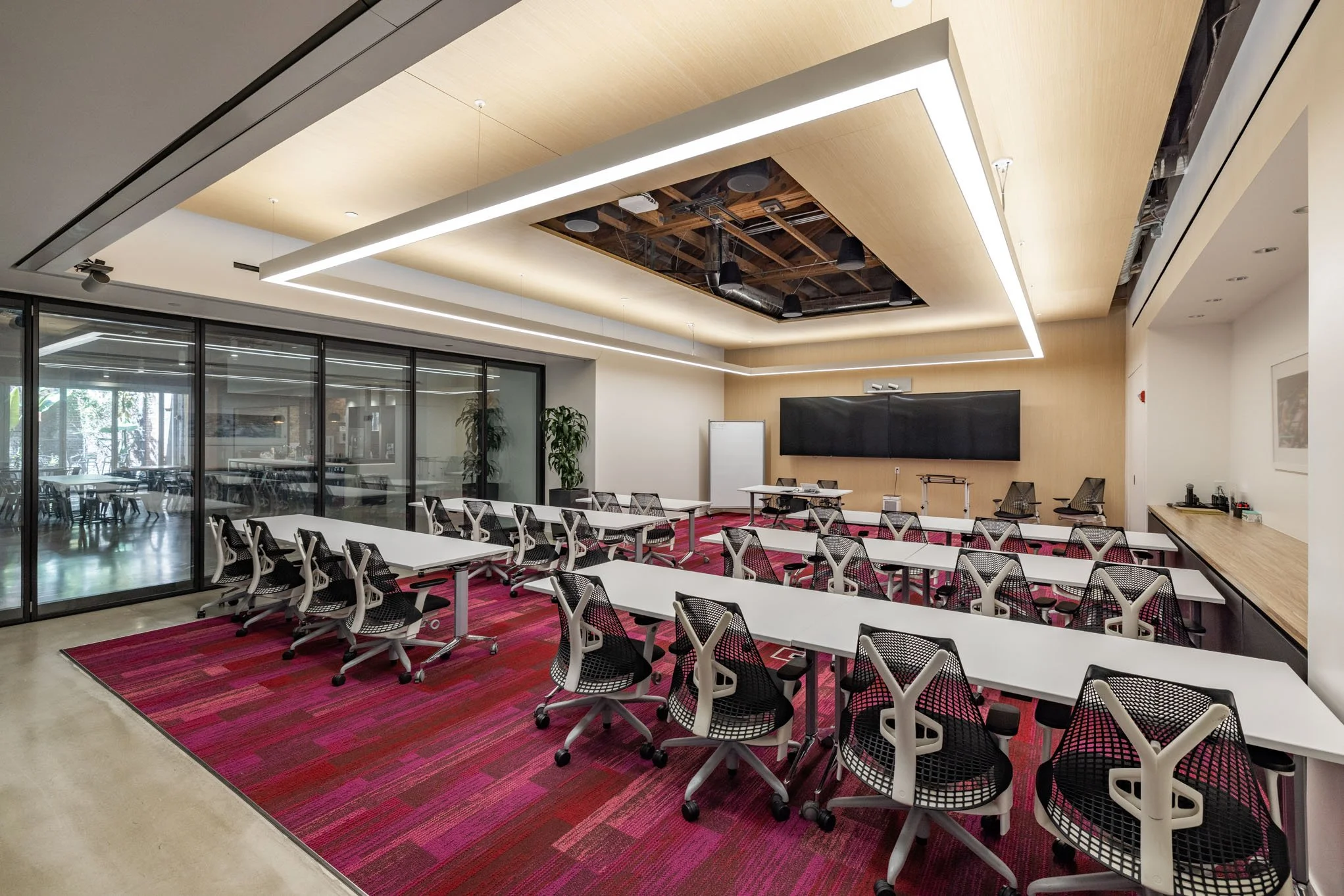 Modern conference room with white tables, black mesh chairs, a large digital screen, a whiteboard, and a windowed partition wall with trees outside. The room has a pink patterned carpet and overhead lighting.