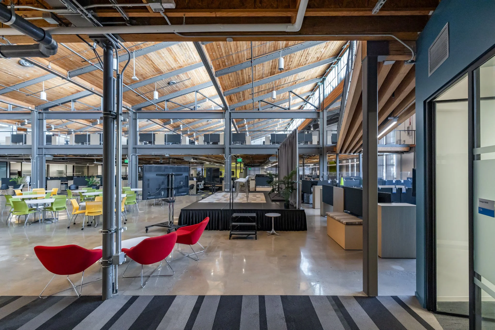 Open office space with a small stage, colorful chairs, and a high ceiling with wood and metal beams.