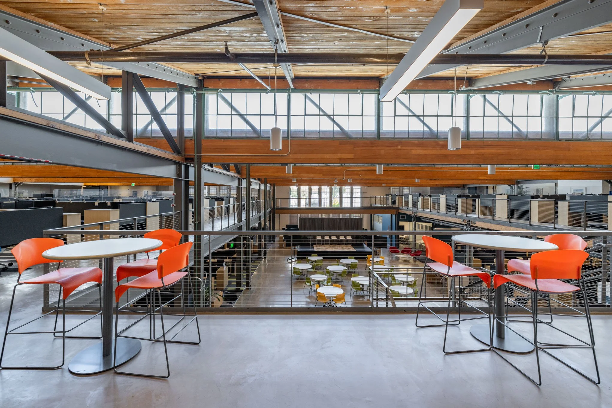 Modern open-concept office space with orange chairs and white tables on a balcony overlookin a ground floor area with colorful chairs and tables.