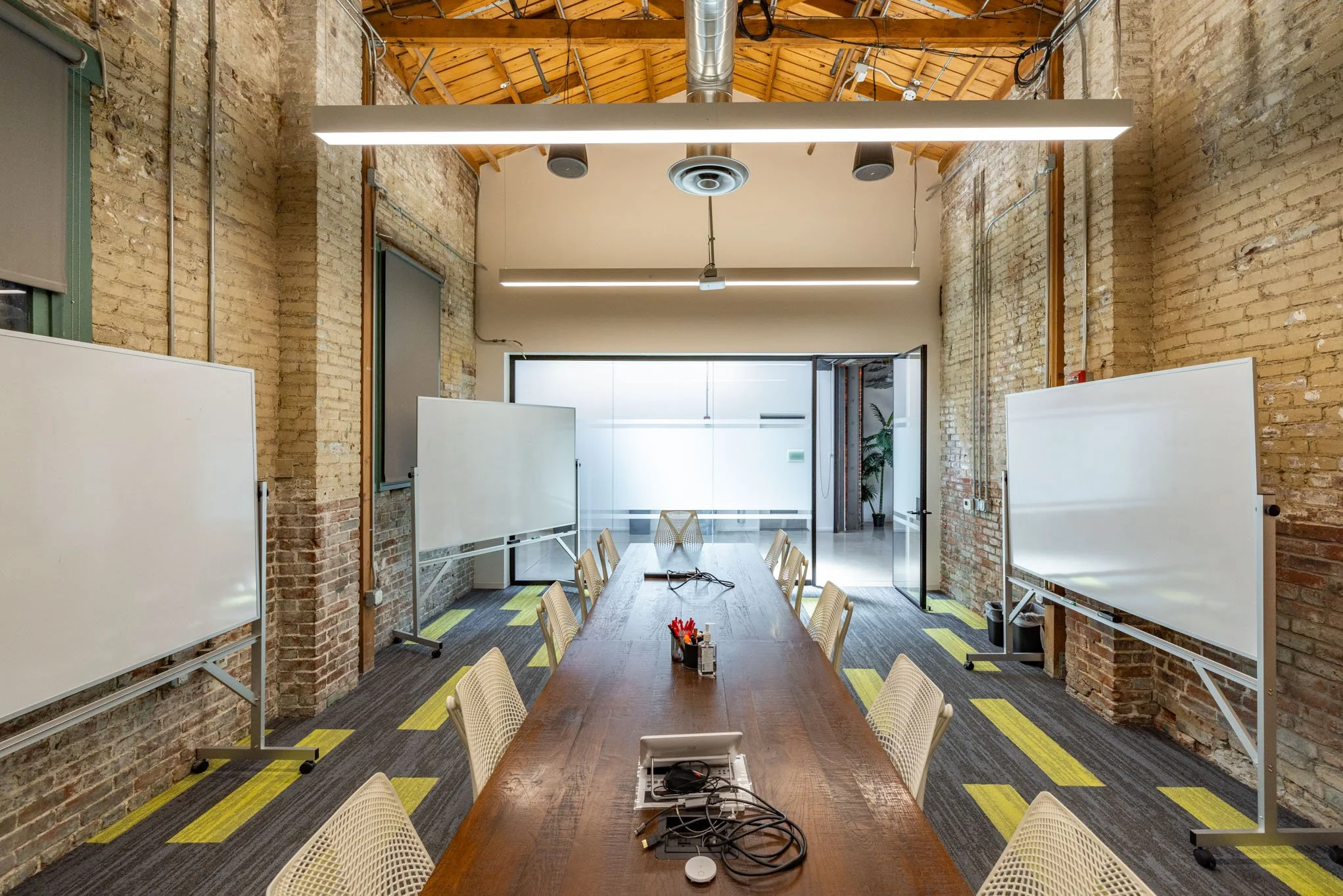 A modern conference room with a long wooden table, white chairs, whiteboards on either side, exposed brick walls, and a high ceiling with wooden beams and industrial lighting. There are glass doors at the back leading to another space with a potted p
