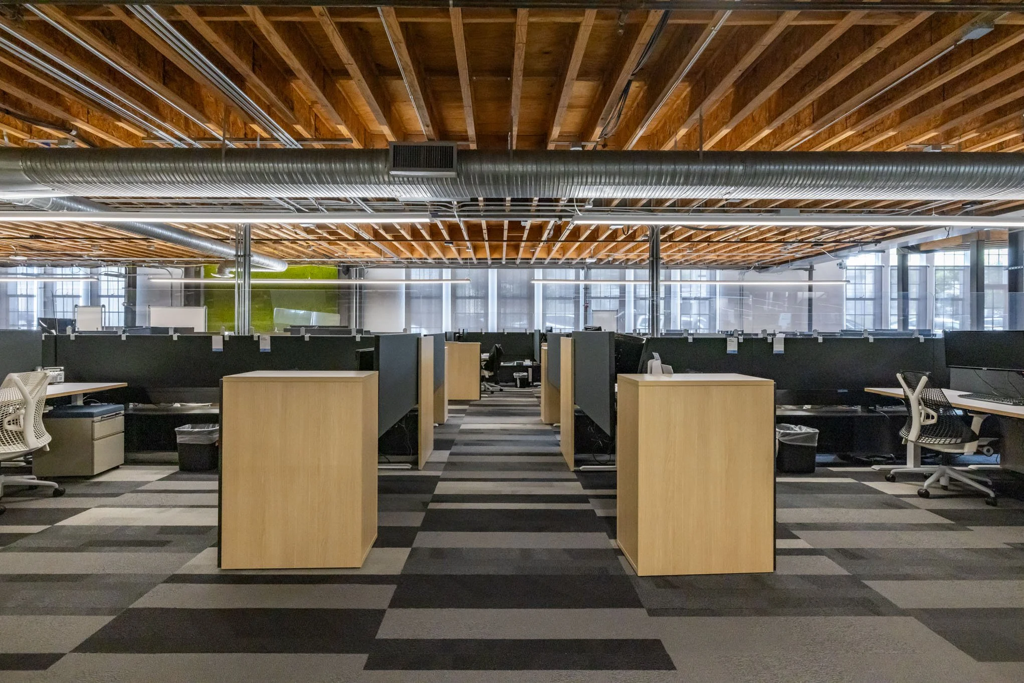 Empty modern office space with cubicles, chairs, and a patterned carpet. Exposed wooden ceiling beams and large windows provide natural light.