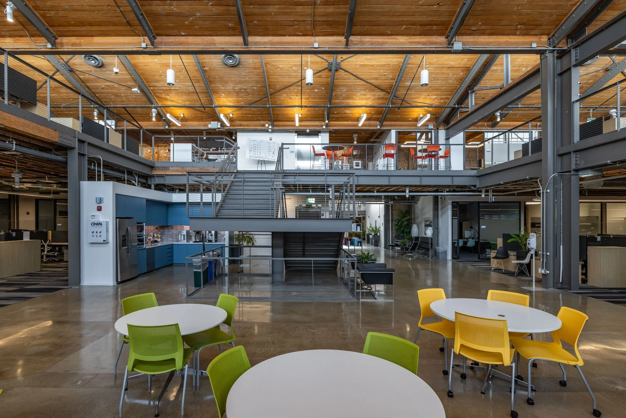 Interior view of a modern office space with a two-level open layout, colorful chairs around white tables in the foreground, metal staircase in the center, and work areas with chairs and desks on both floors under a wooden ceiling.