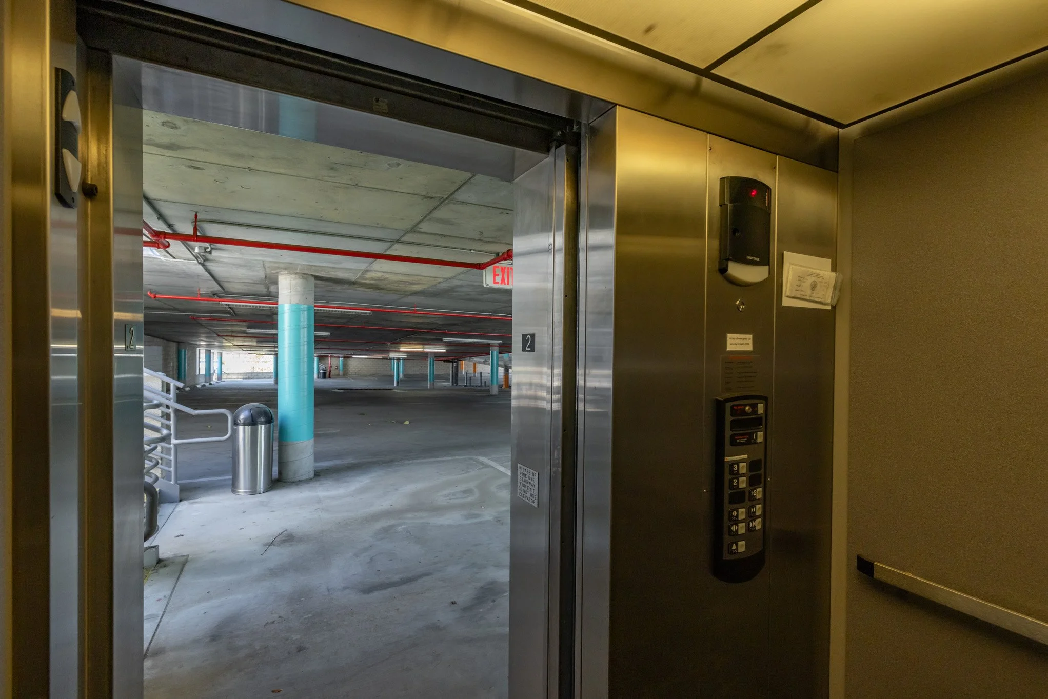 Elevator door looking out into a multi-level parking garage with blue-painted columns, a trash can, and parking space markings.