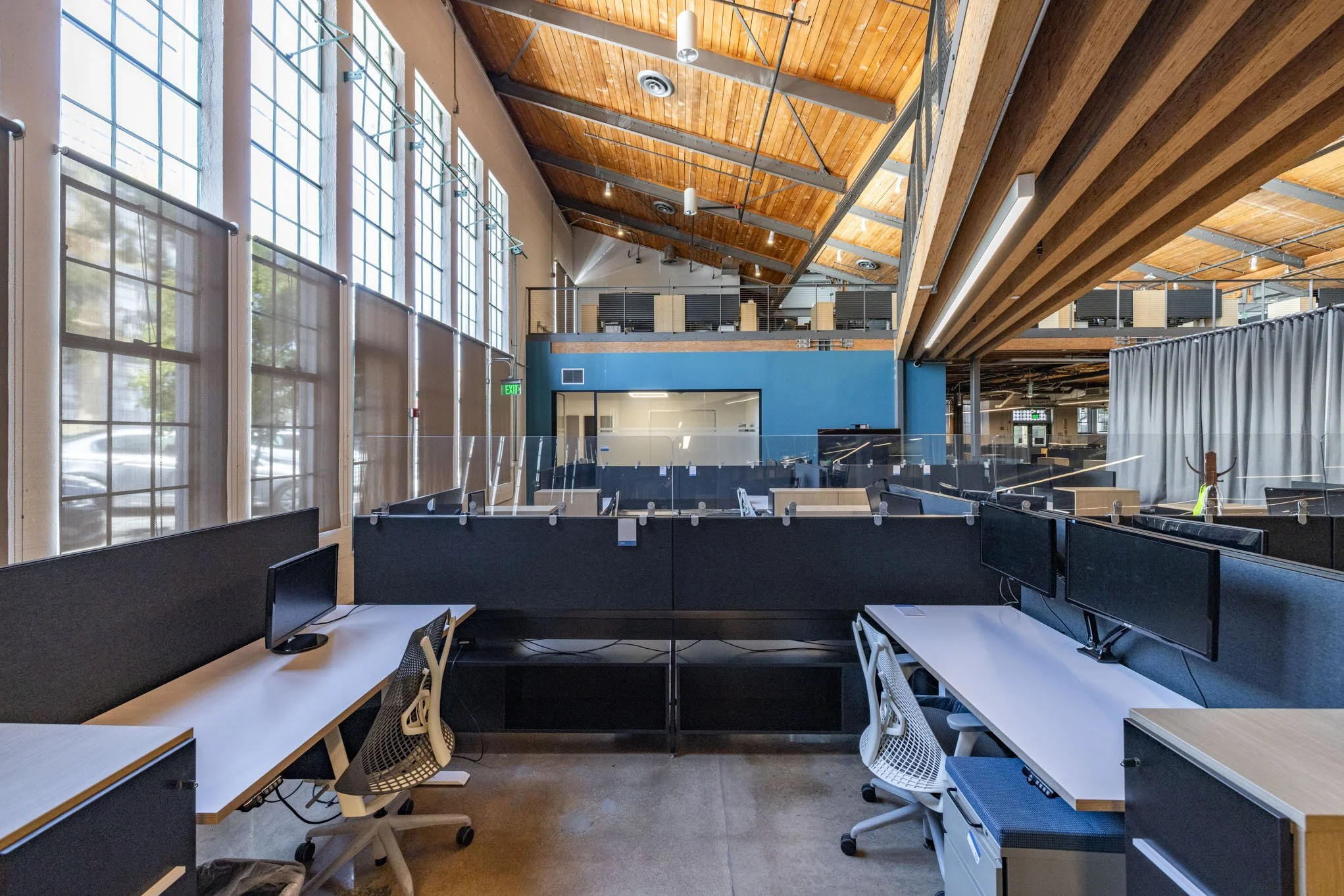 Modern open-concept office space with workstations, computers, and high ceilings with exposed wood beams and large windows.