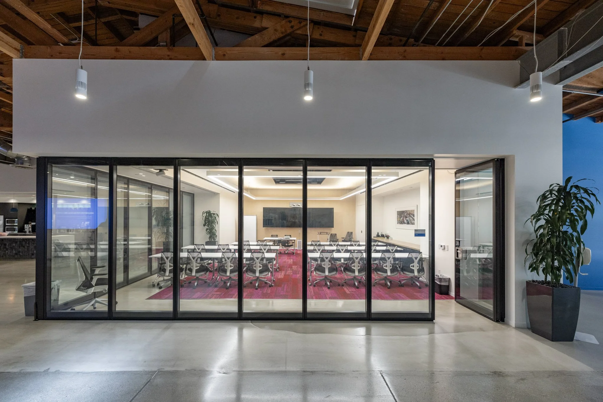 Modern conference room with glass walls, equipped with multiple chairs and a large screen on the wall, inside a building with exposed wooden beams and a polished concrete floor.