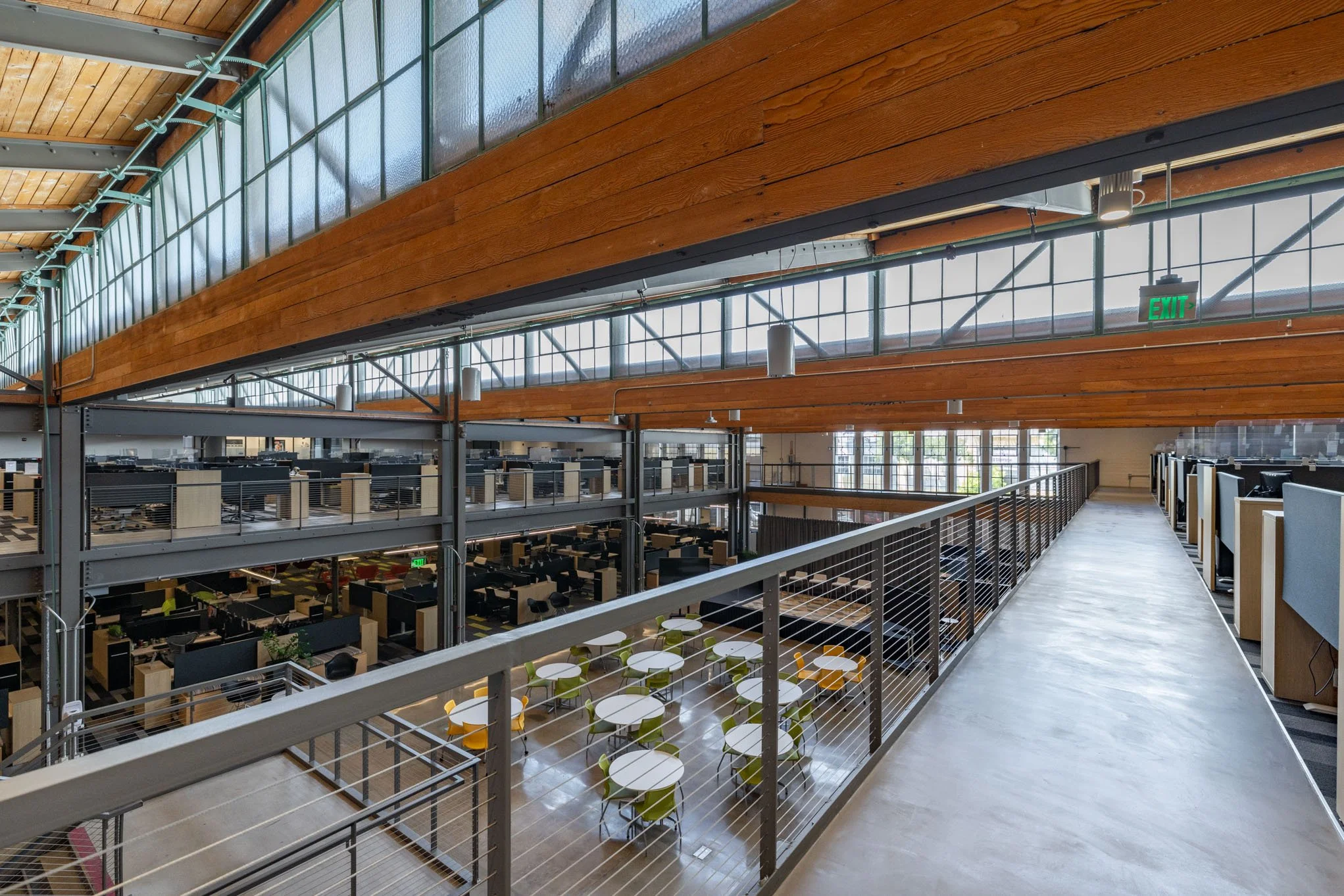 Modern office workspace with multiple floors, glass walls, wooden ceiling accents, and a balcony with tables and chairs.