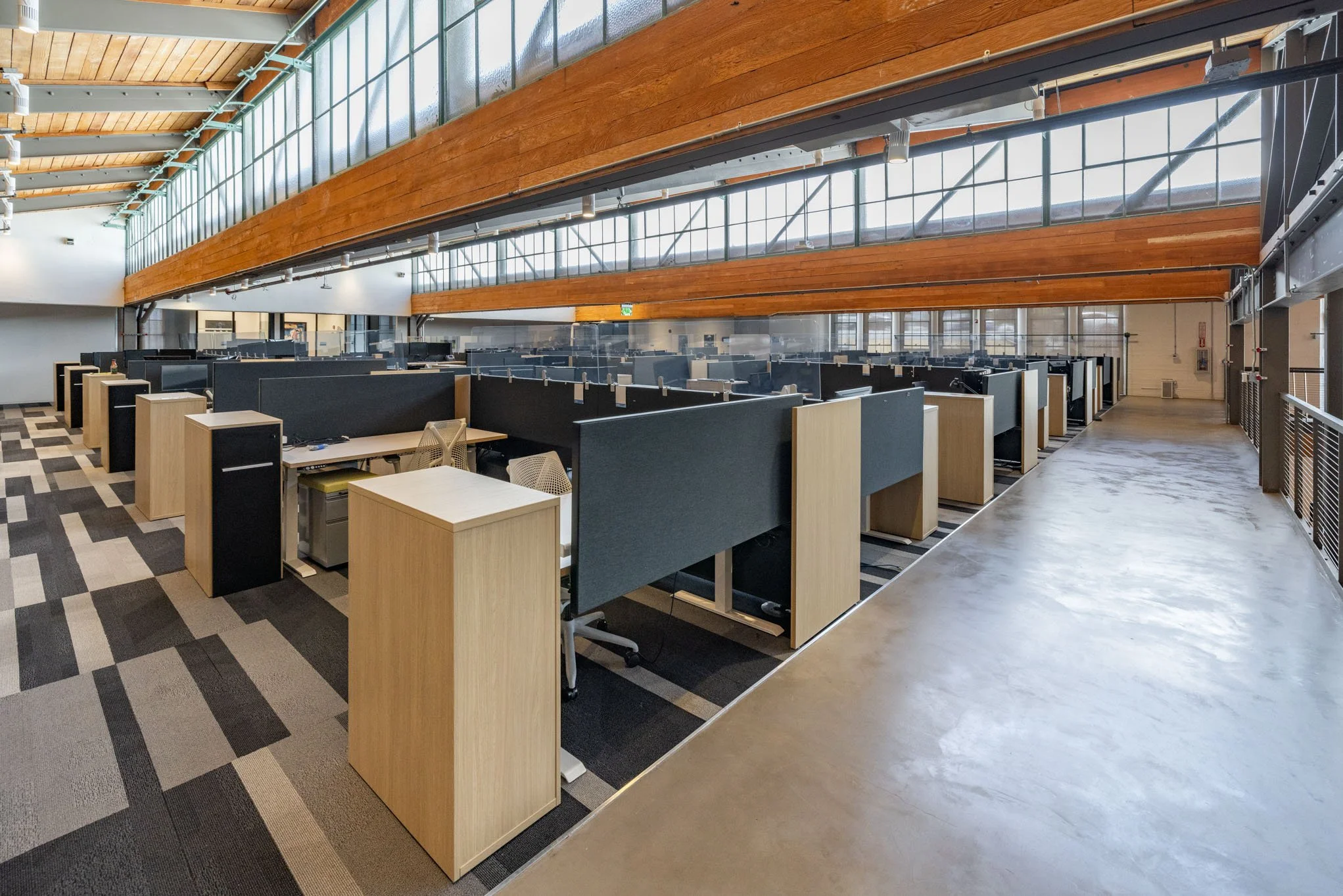 Empty office cubicles in a modern, well-lit workspace with large windows, wood ceiling beams, and a patterned carpet.