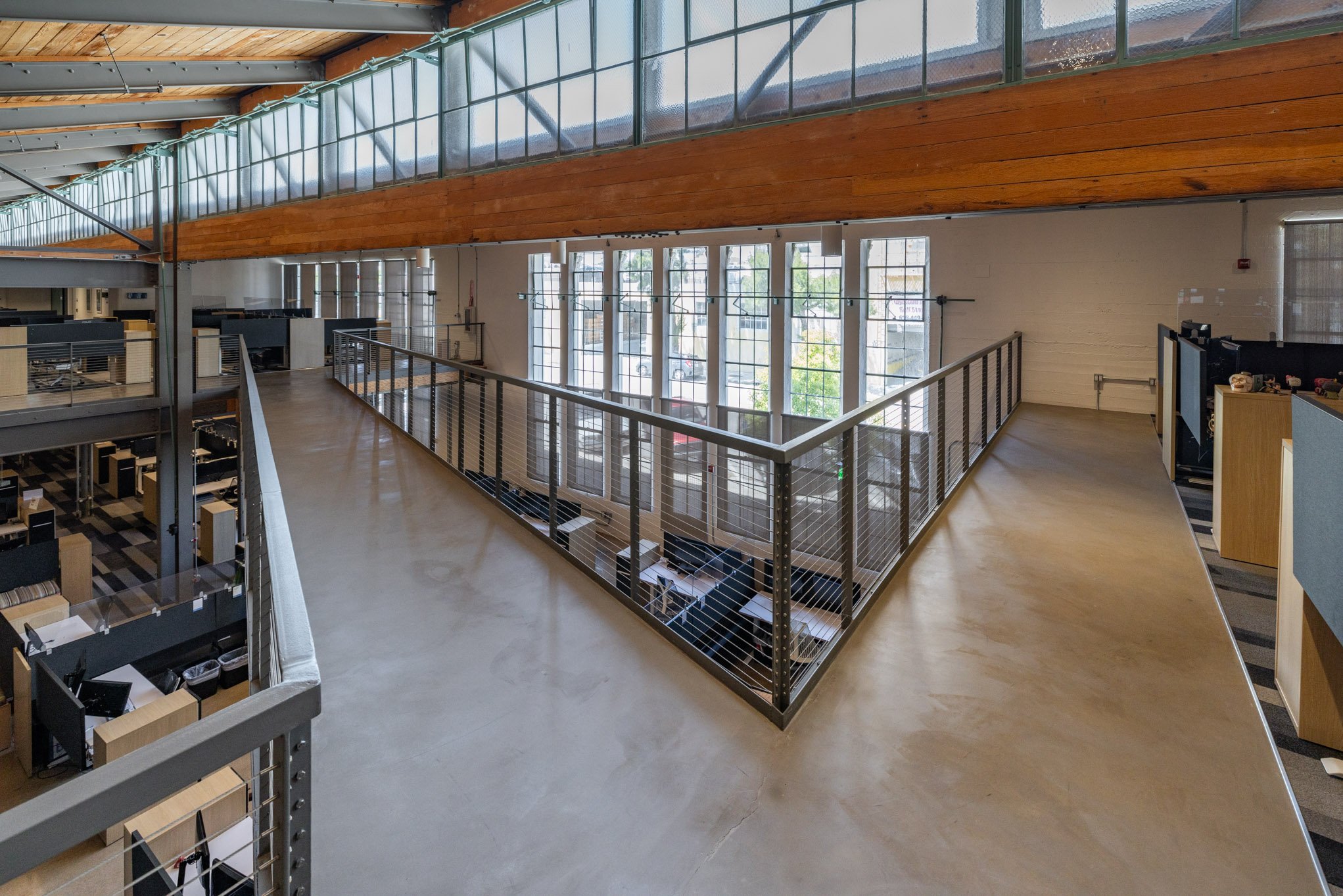Interior view of a modern office space with a mezzanine level, large windows, and wooden ceiling beams.