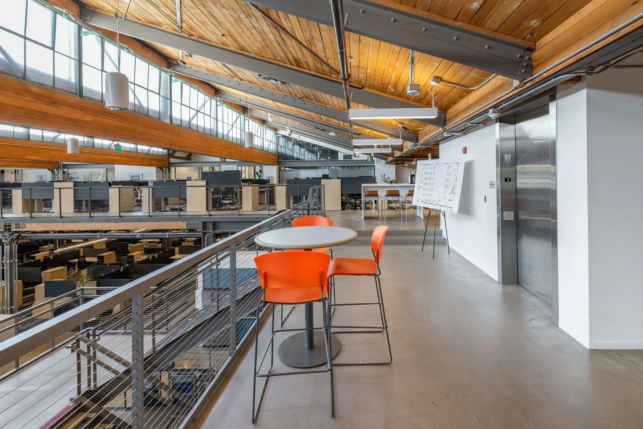 Modern office space with open layout, wooden ceiling, large windows, and a balcony with a round table and orange chairs.