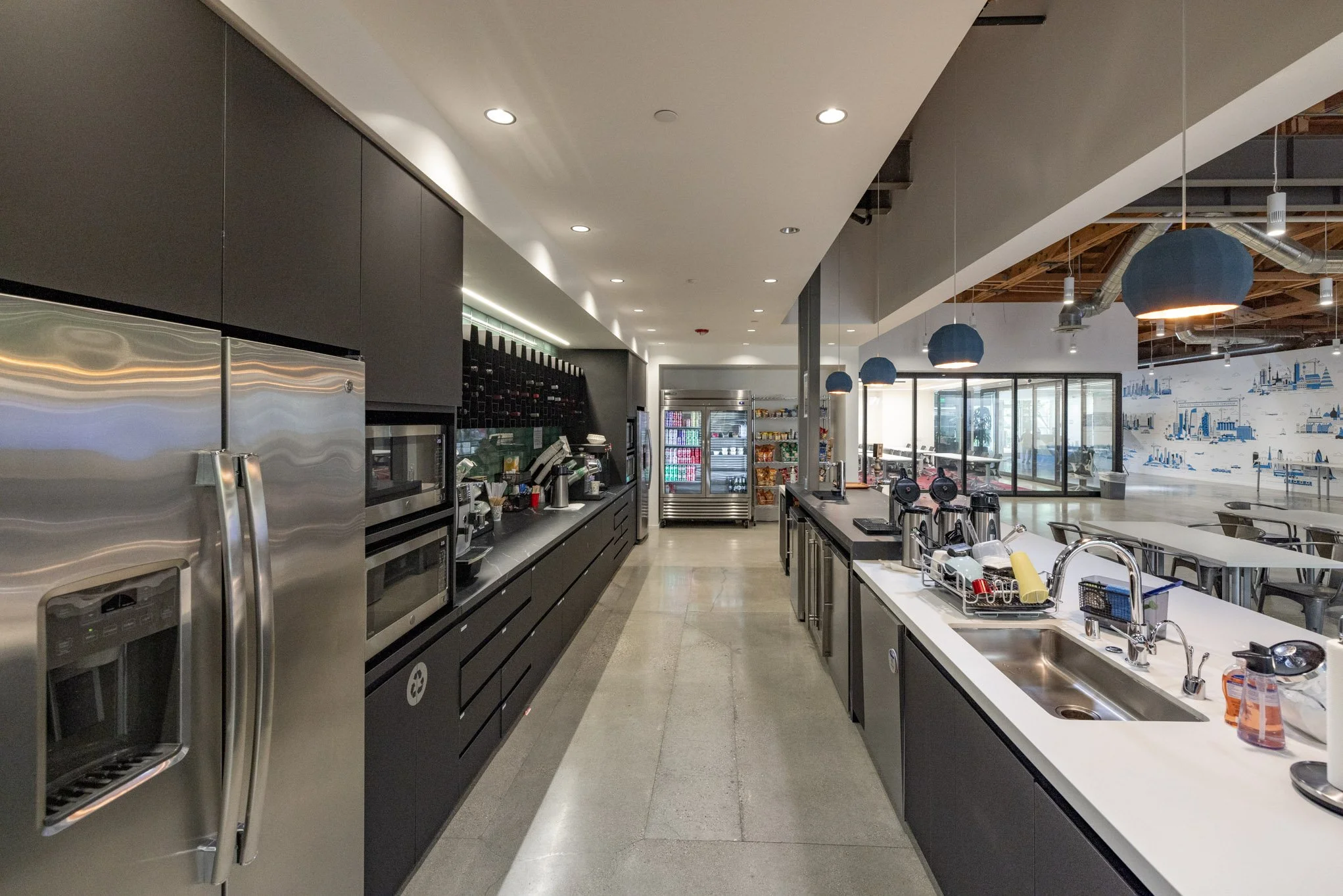 Modern kitchen area with stainless steel appliances, black cabinets, and a white countertop with various kitchen items, in an open-concept space with a seating area and large windows in the background.