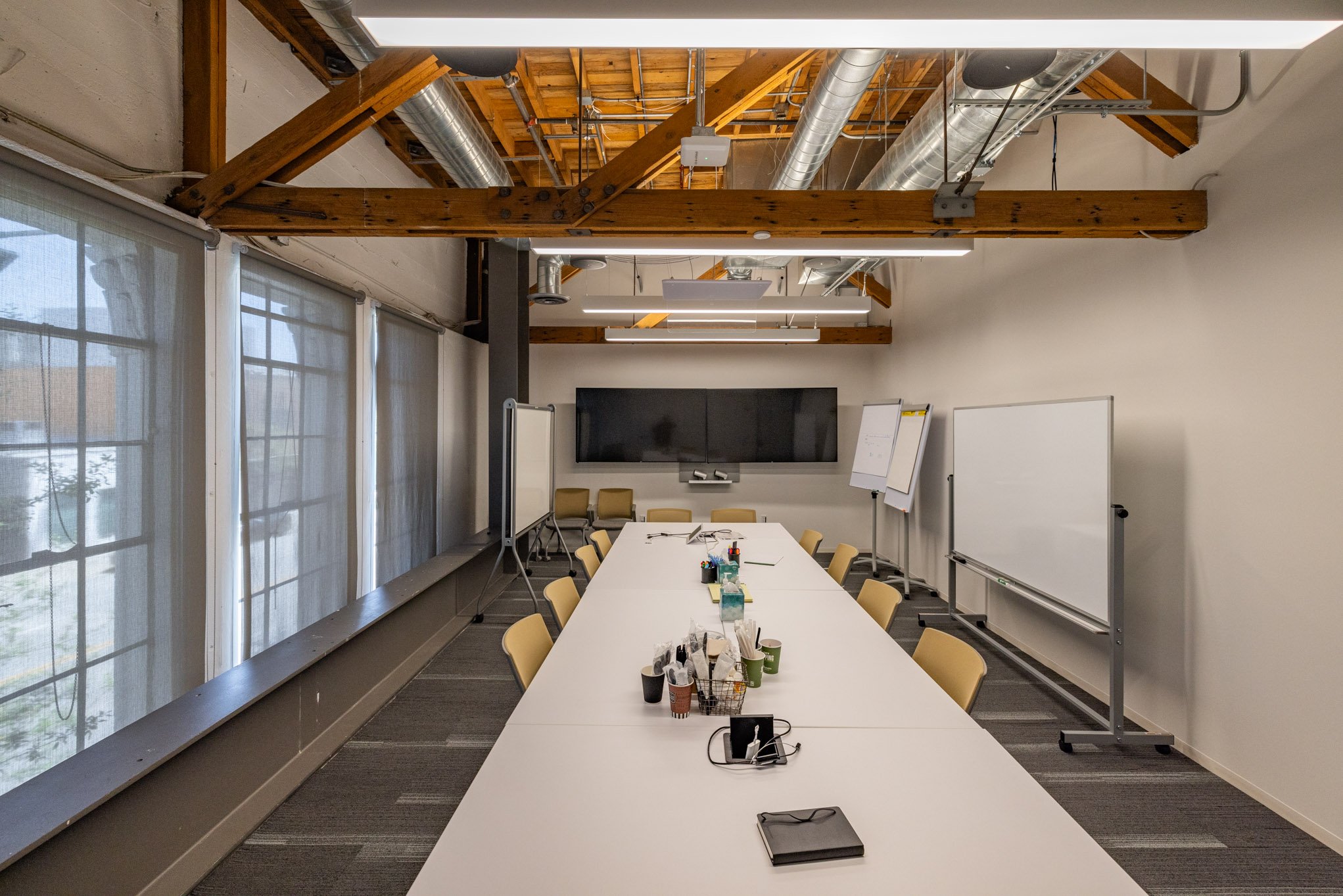An empty conference room with a long table, chairs, whiteboards, a large screen, and windows with blinds.