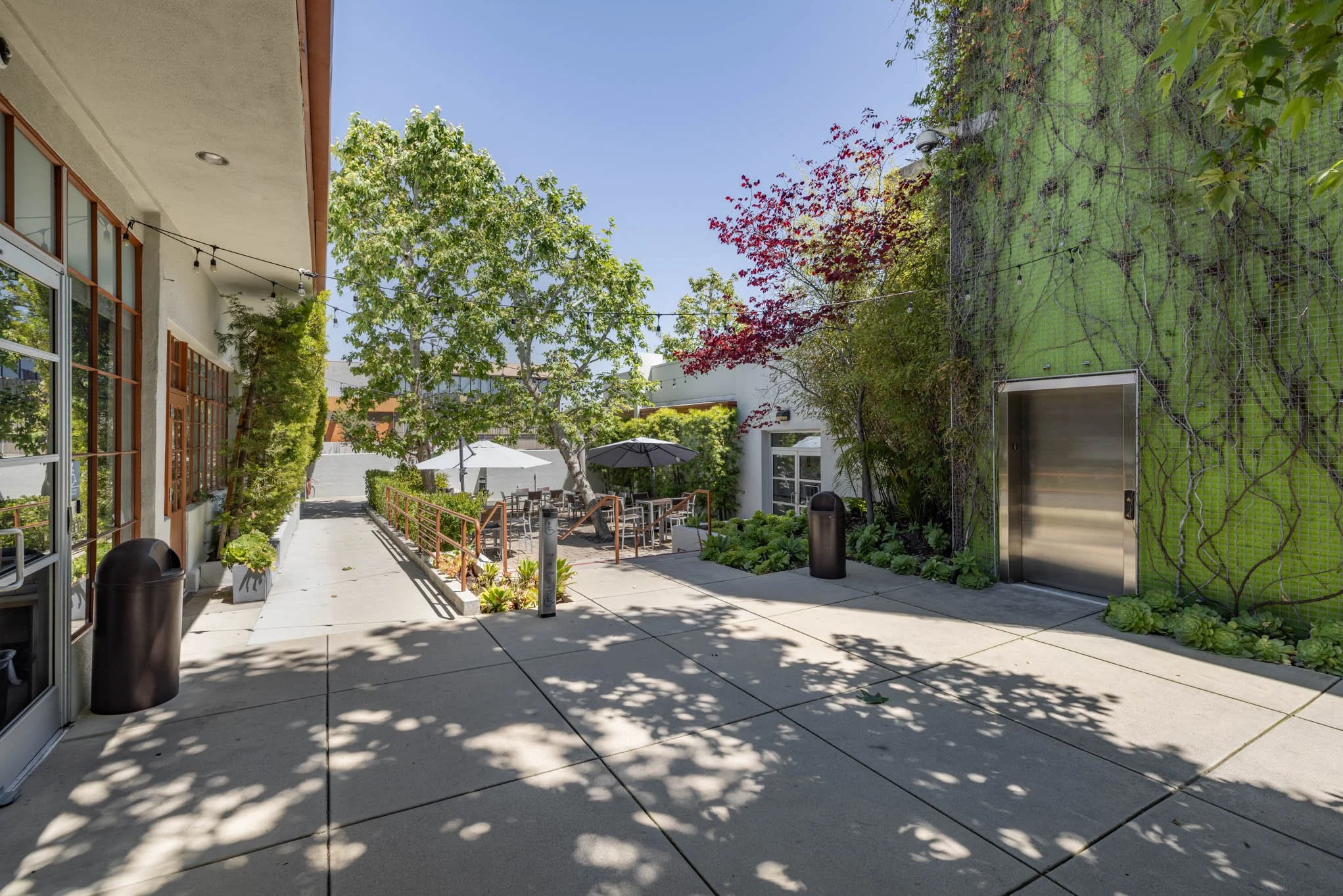 An outdoor patio area with tables, chairs, umbrellas, trees, and a green wall with an elevator.