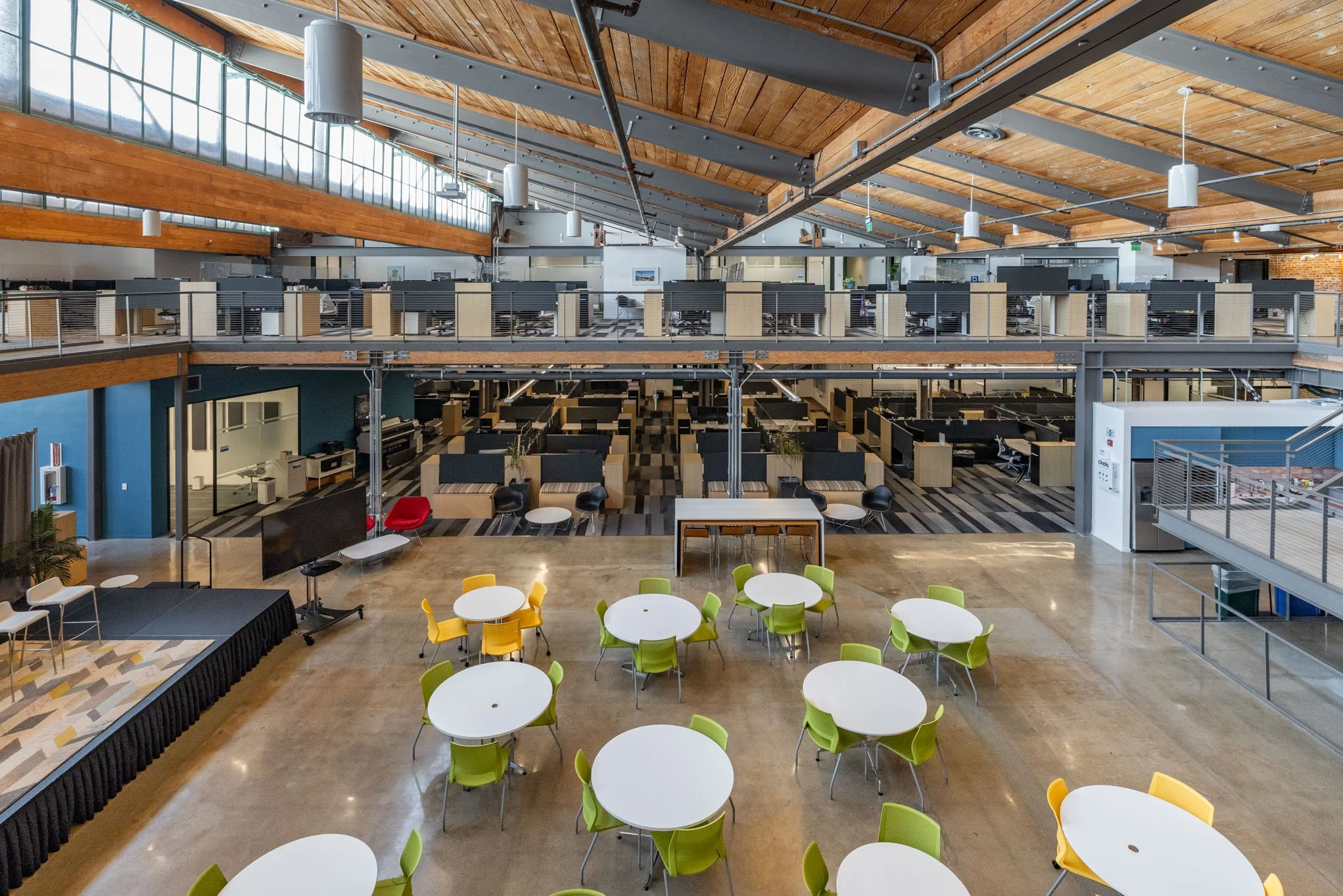 Interior view of a modern, open-concept office space with a high wooden ceiling, large windows, and multiple workstations on upper and lower levels. There are round tables with green and yellow chairs in the foreground, and a lounge area with a TV, r