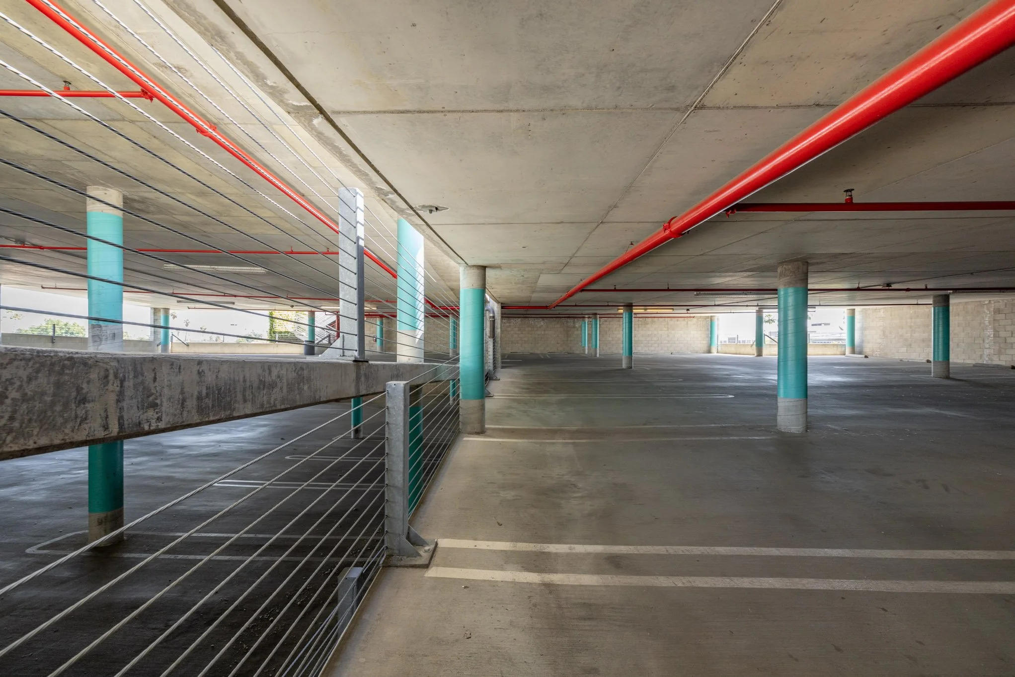 Empty multi-level parking garage with concrete floor, support columns wrapped in teal protective covering, red fire sprinkler pipes, and metal safety barriers.