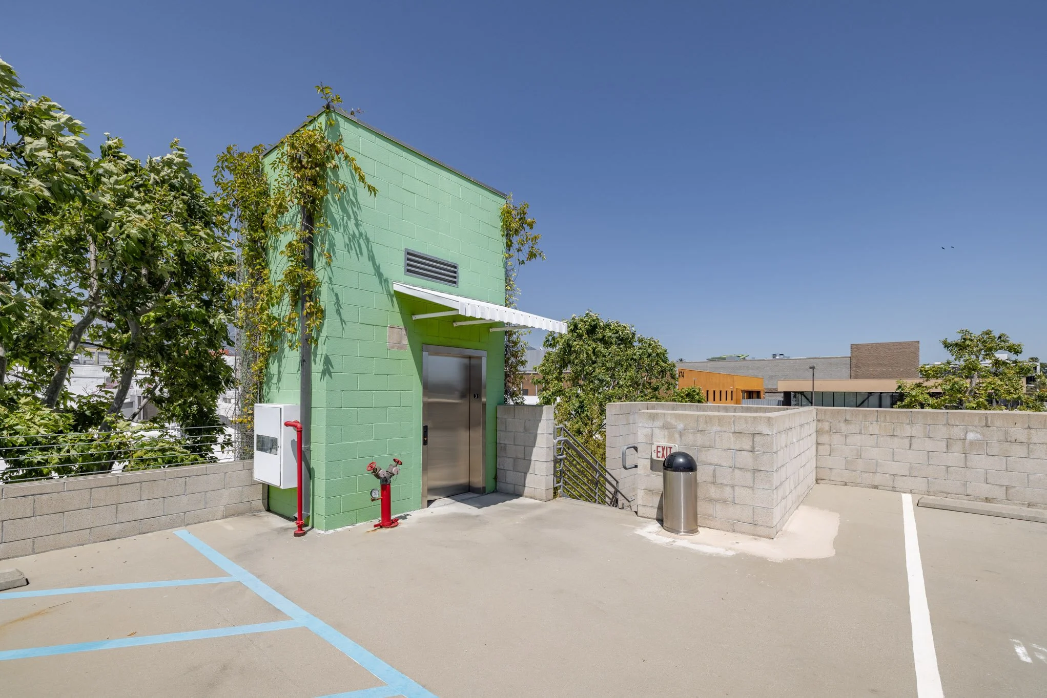 Rooftop parking area with a small green building, trees, a trash can, and parking lines under a clear blue sky