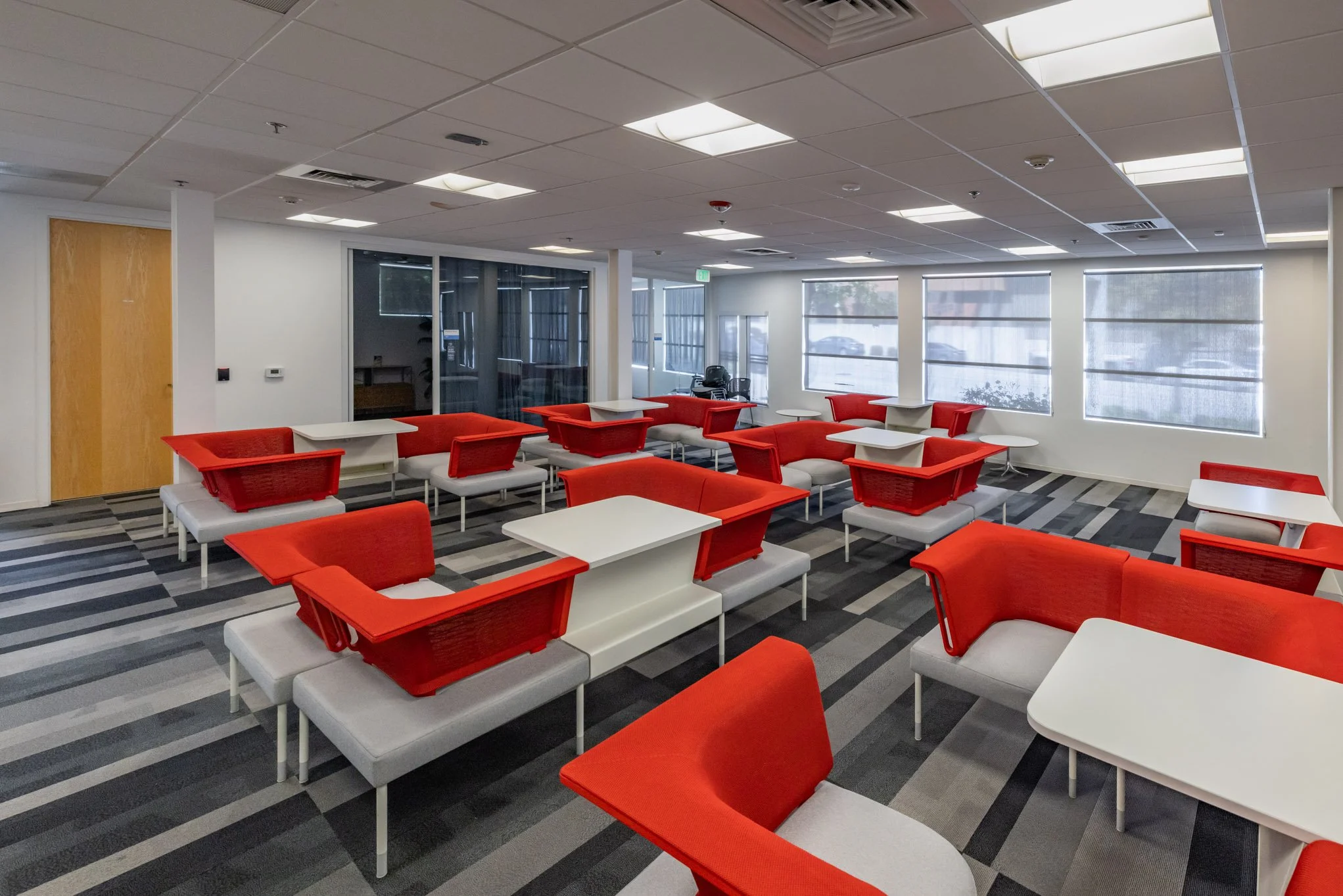 Empty waiting area with white tables, red and gray chairs, and large windows with blinds.