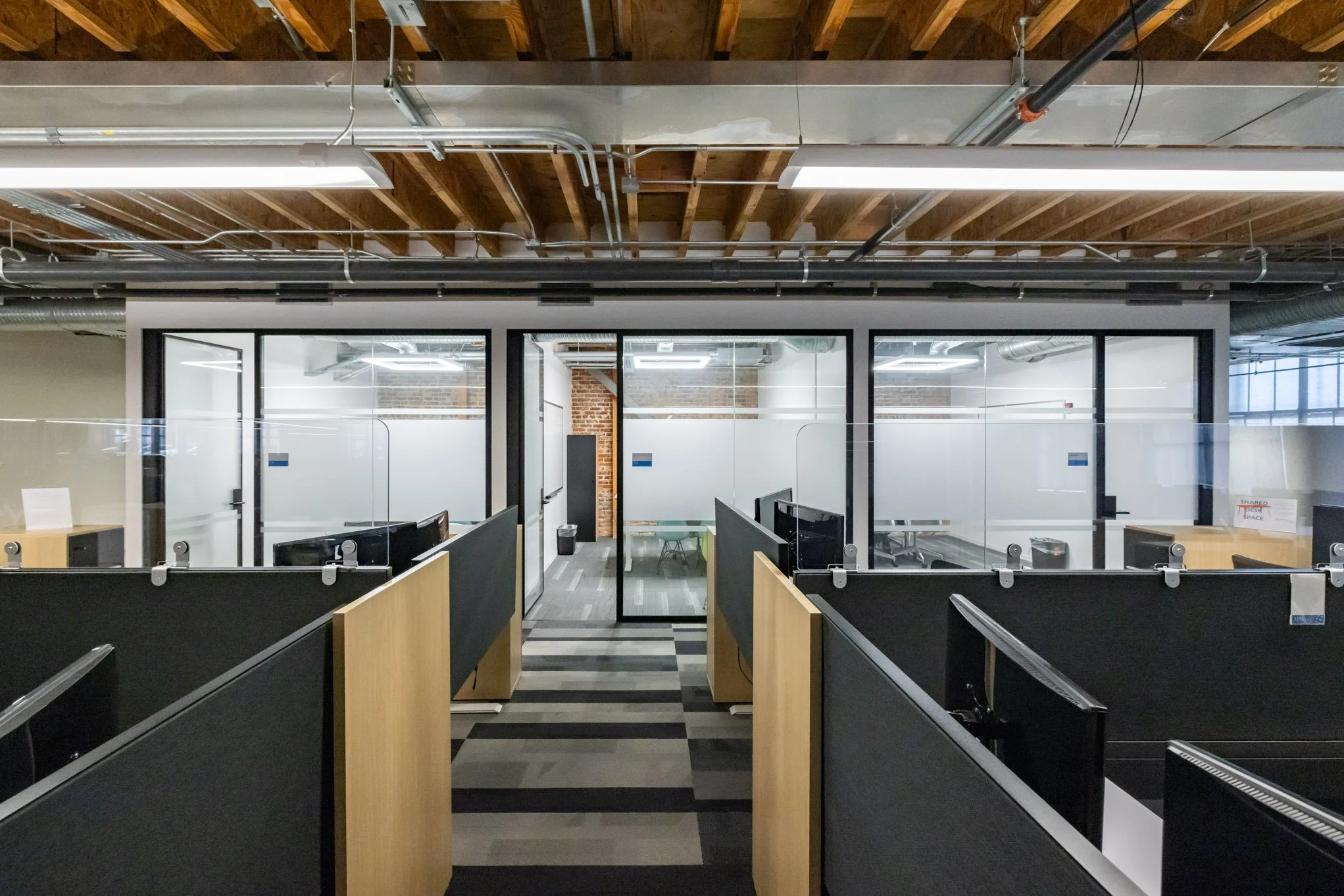 An empty modern office space with cubicles, glass partitions, and fluorescent lighting, featuring wood ceiling beams and exposed ductwork.