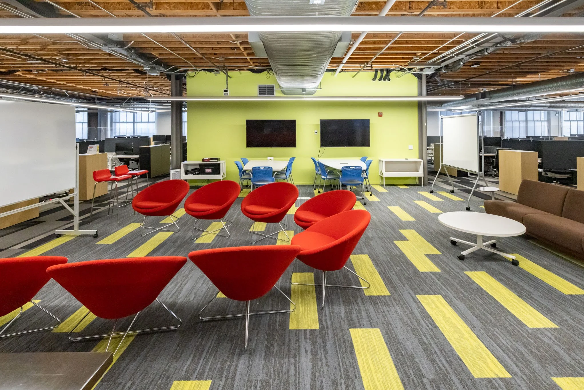 An office space with red modern chairs, a brown sofa, white tables, two black wall-mounted screens, whiteboards, and workstations with computers. The room has a green accent wall and a ceiling with exposed wood and ductwork.