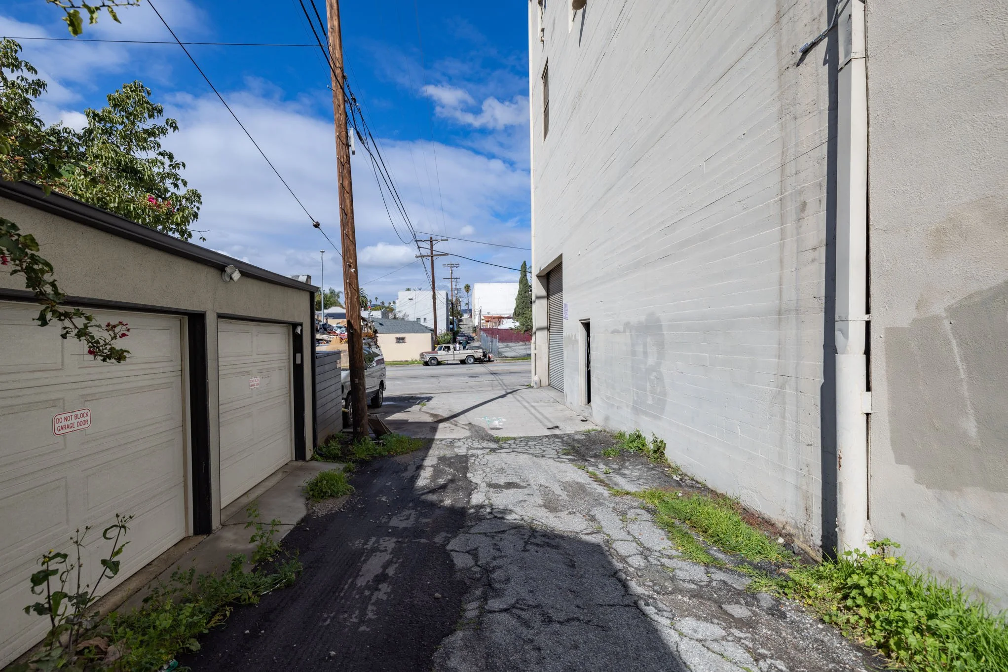 An alleyway with a cracked pavement, a utility pole, a white brick wall on the right, and a building with a garage door on the left, under a partly cloudy blue sky.