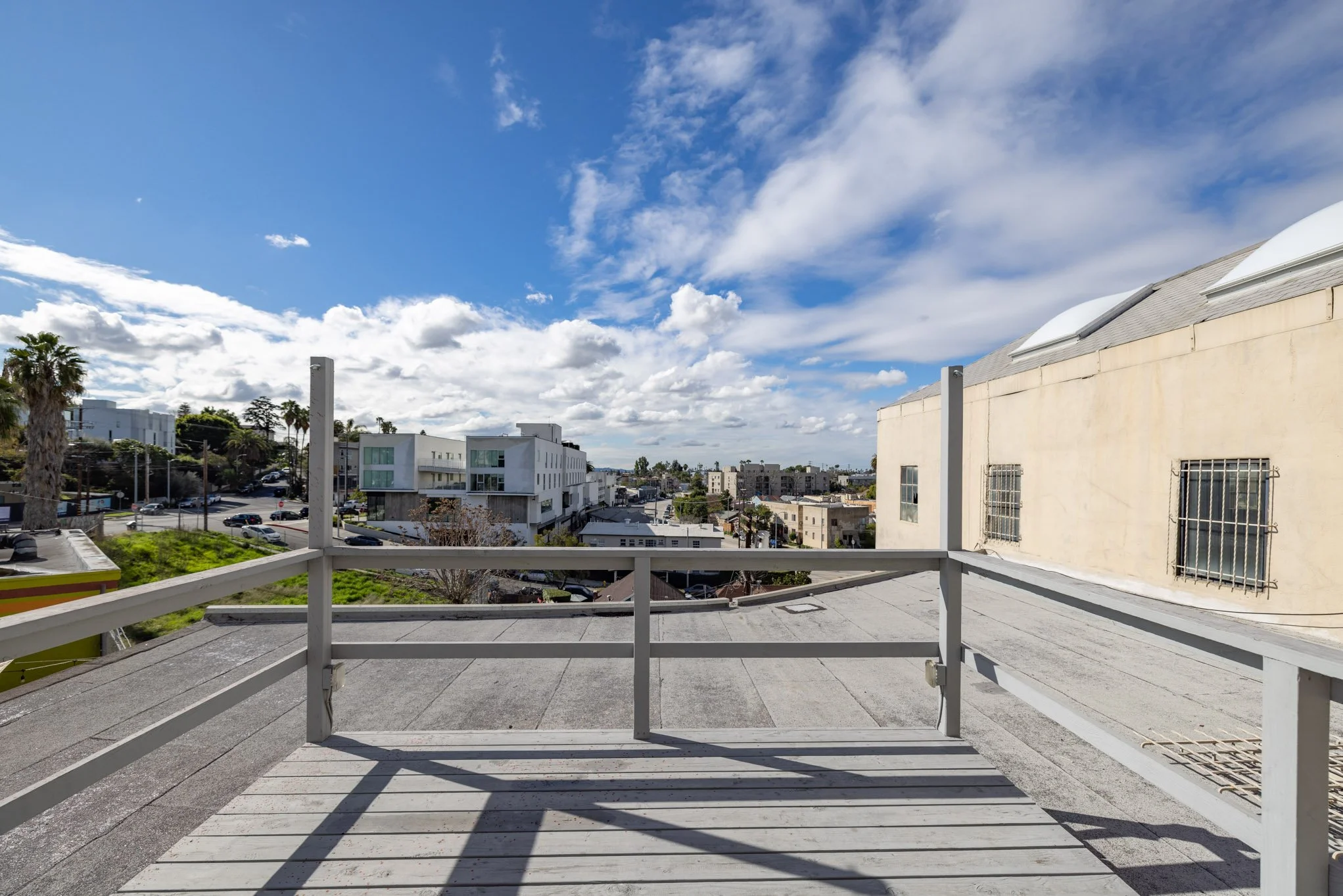 View from a rooftop balcony overlooking a cityscape with modern buildings, parking lots, trees, and a partly cloudy blue sky.