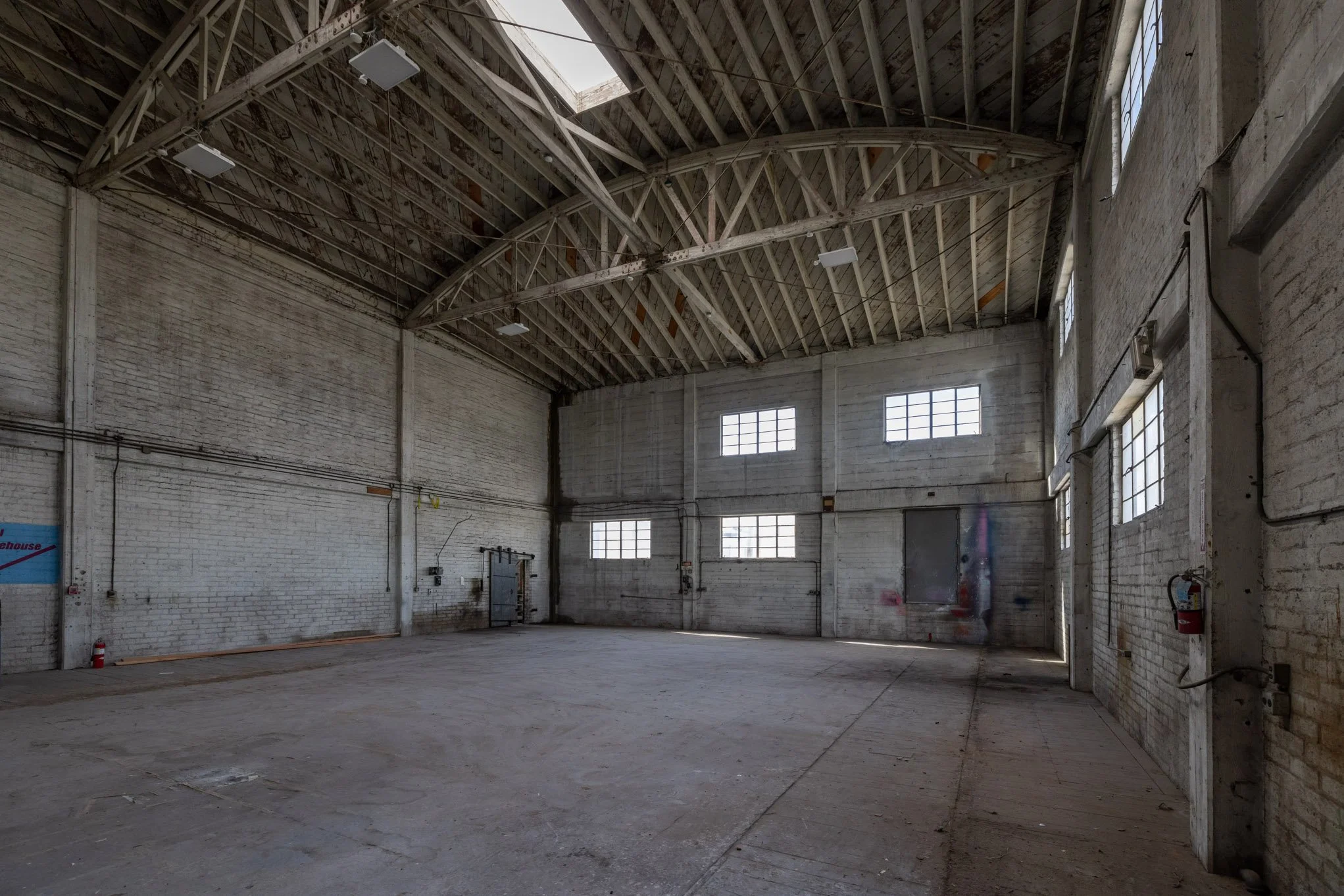 Interior of an empty industrial warehouse with high ceilings, exposed beams, white brick walls, and several windows providing natural light.