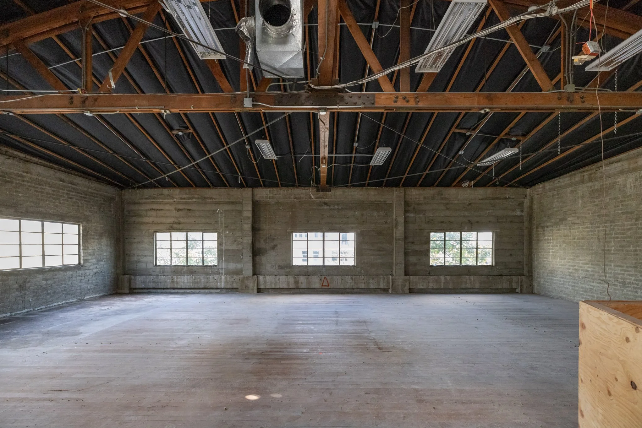 Empty industrial-style room with wooden floor, exposed brick walls, and large windows, under a high ceiling with visible wooden beams and ductwork.