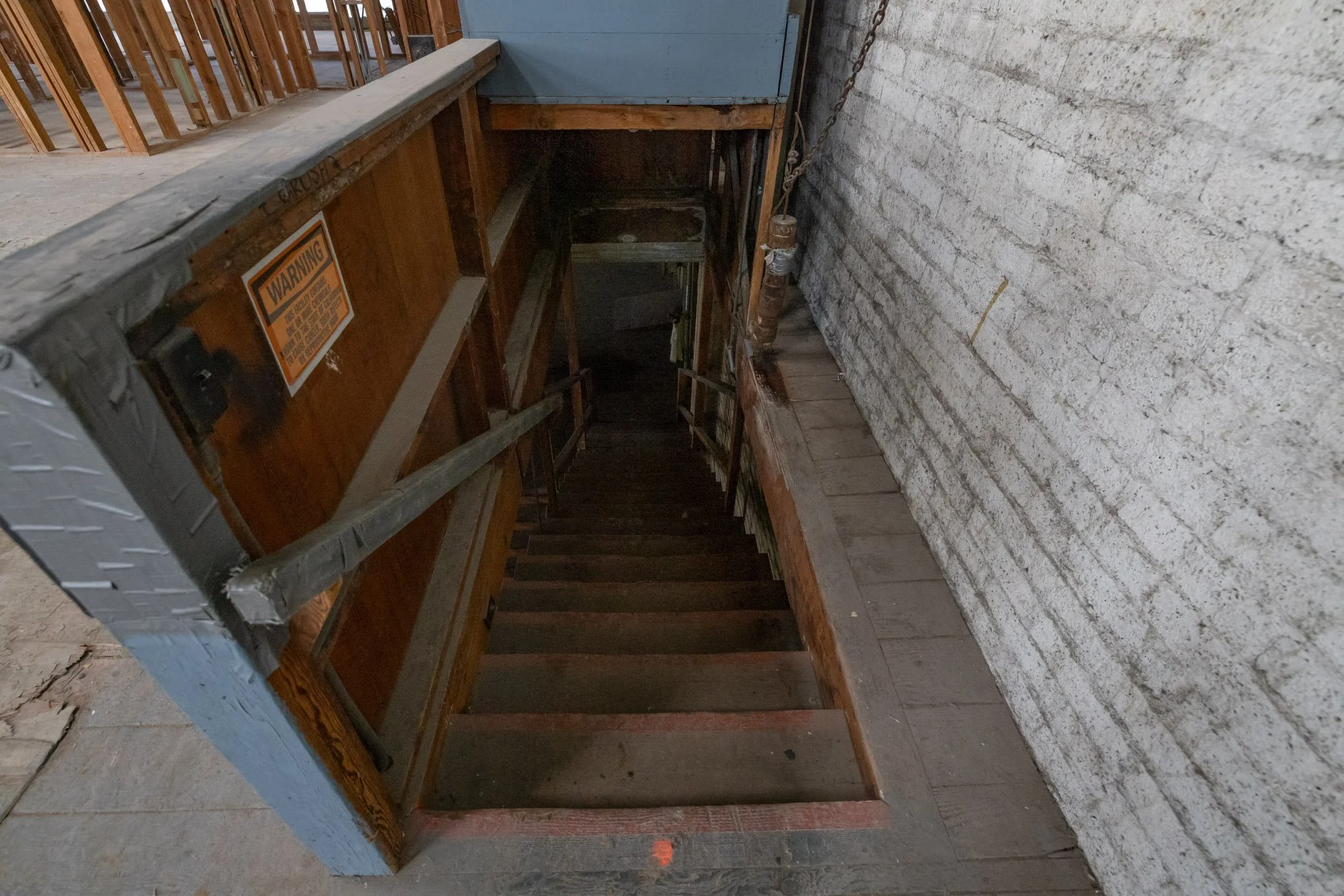 Indoor staircase leading downward with wooden steps, a brick wall on the right, and a wooden railing on the left. A warning sign is visible on the railing.