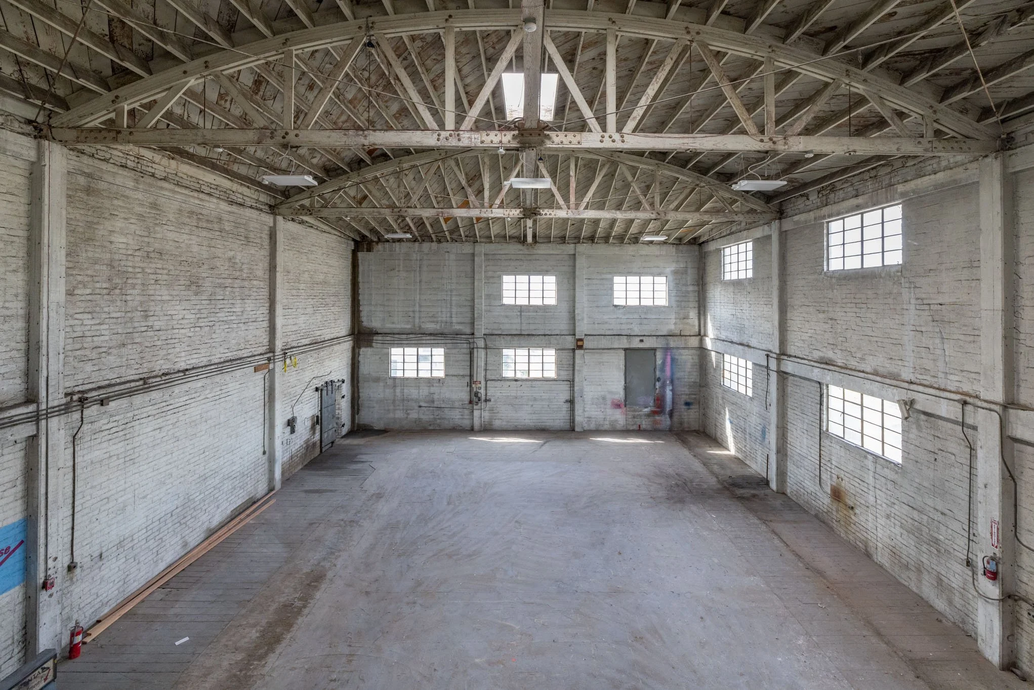 Empty industrial warehouse with white brick walls, high ceiling with exposed wooden beams, and multiple windows allowing natural light.