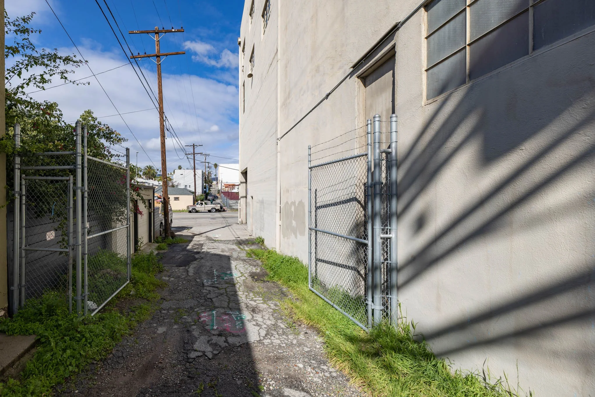 An alleyway with cracked pavement, green grass on the sides, a chain-link fence on the left, and a large white building on the right. Power lines extend overhead, and in the distance, there are trees and parked cars under a partly cloudy sky.