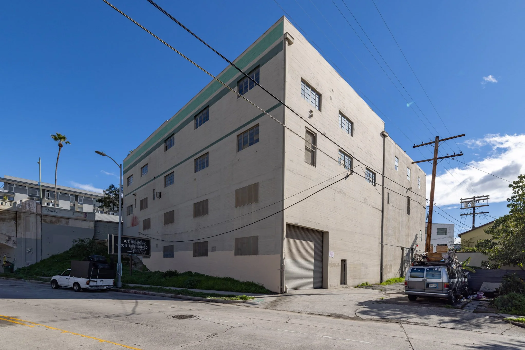 A large, white, multi-story building with small windows, situated on a street corner under a clear blue sky. There are utility poles and wires, a couple of parked cars, and a palm tree in the background.
