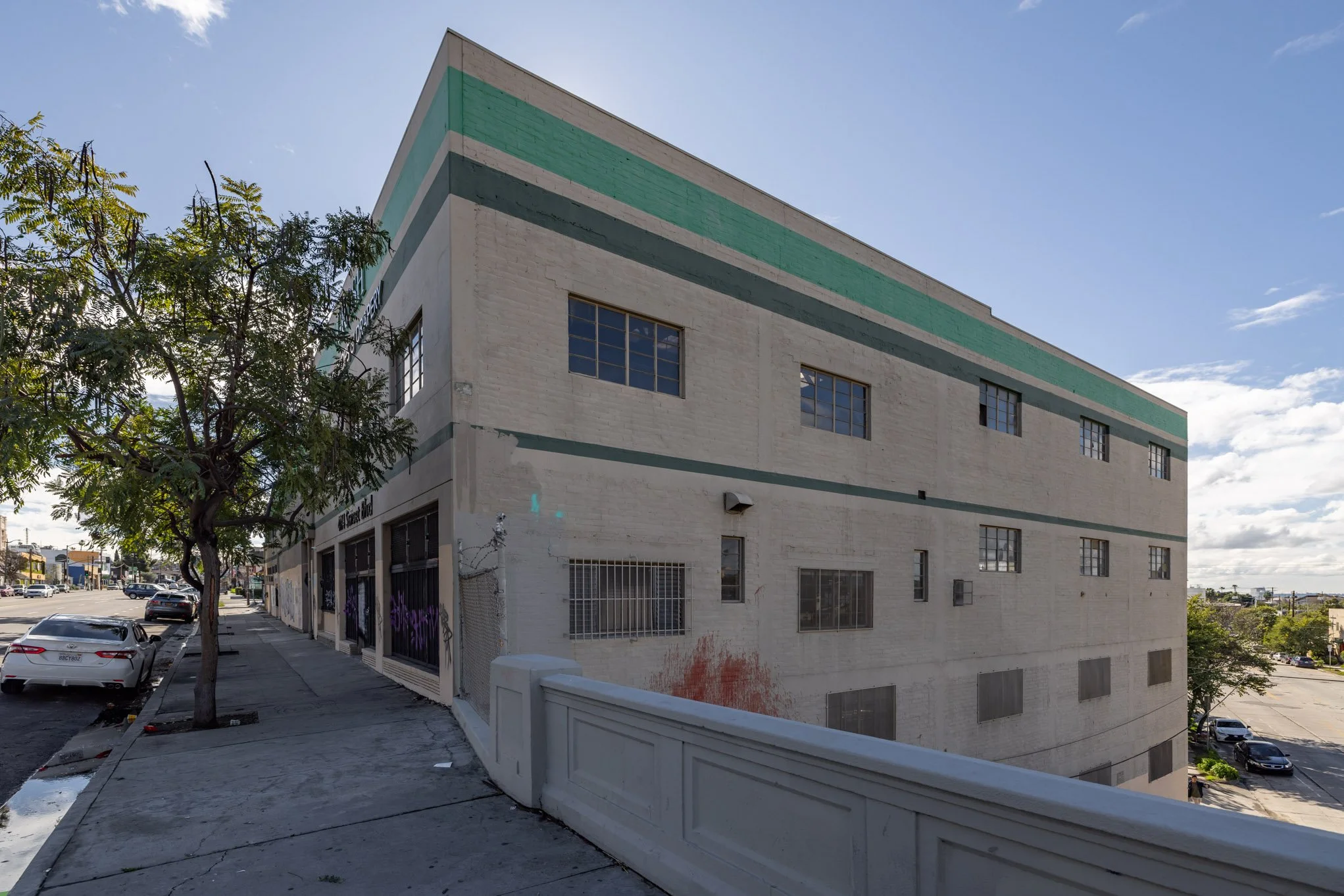 A beige, three-story building with green horizontal stripes, windows with bars, graffiti, and a sidewalk with parked cars and trees.
