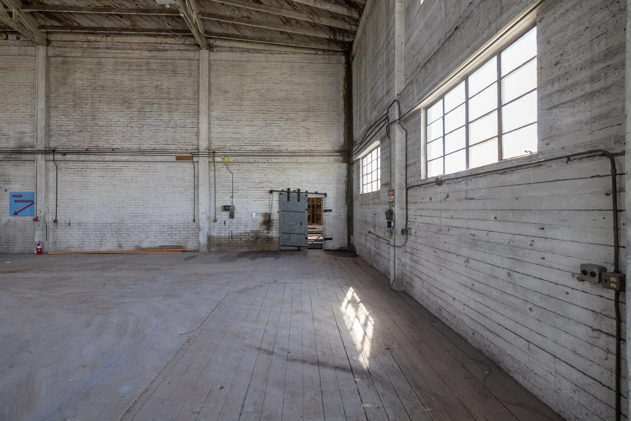 Empty industrial warehouse with white brick and wooden walls, large windows, and a sliding metal door.