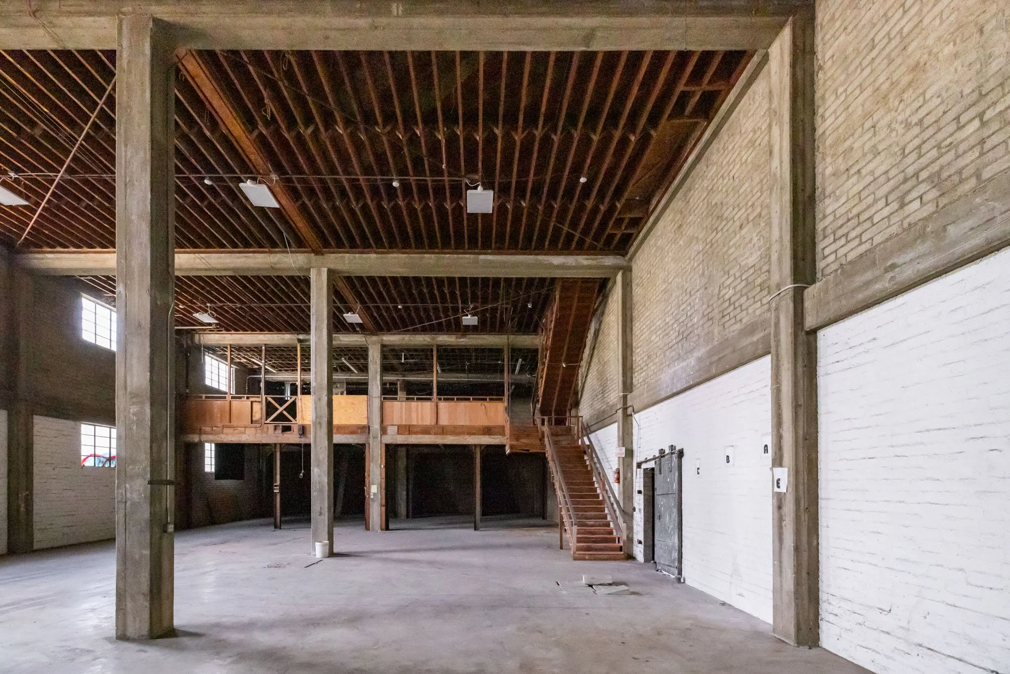 Empty industrial-style warehouse with exposed brick walls, wooden beams, a staircase leading to an upper level, and large windows letting in natural light.