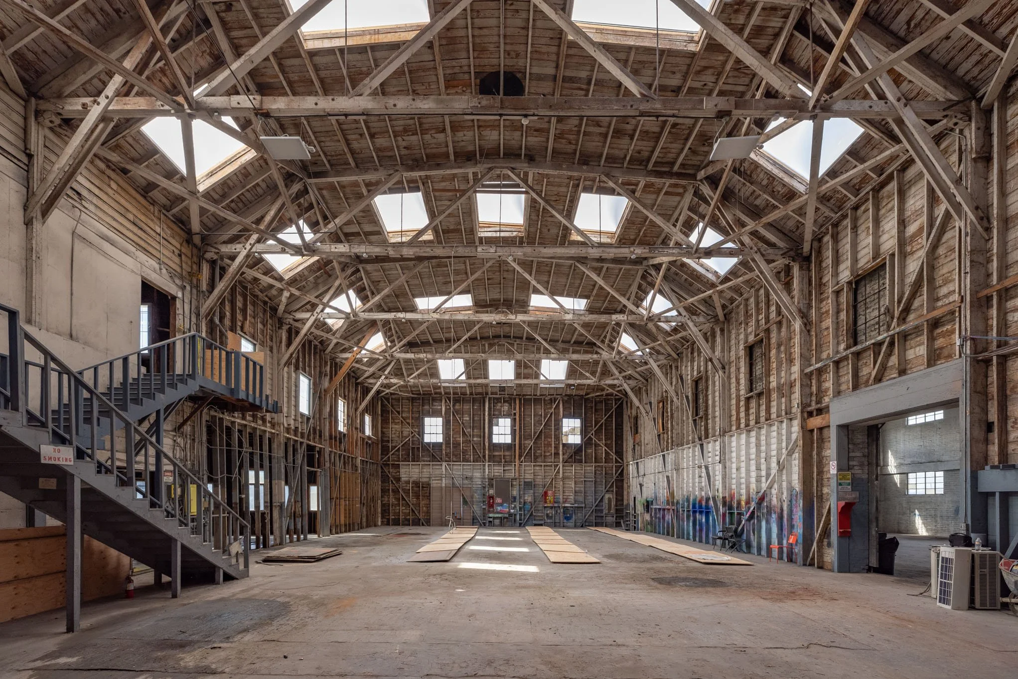 Empty large industrial warehouse with wooden walls and ceiling, skylights, and a mechanical staircase on the left. The floor is bare, and the walls have some pathwork and graffiti on the right side.