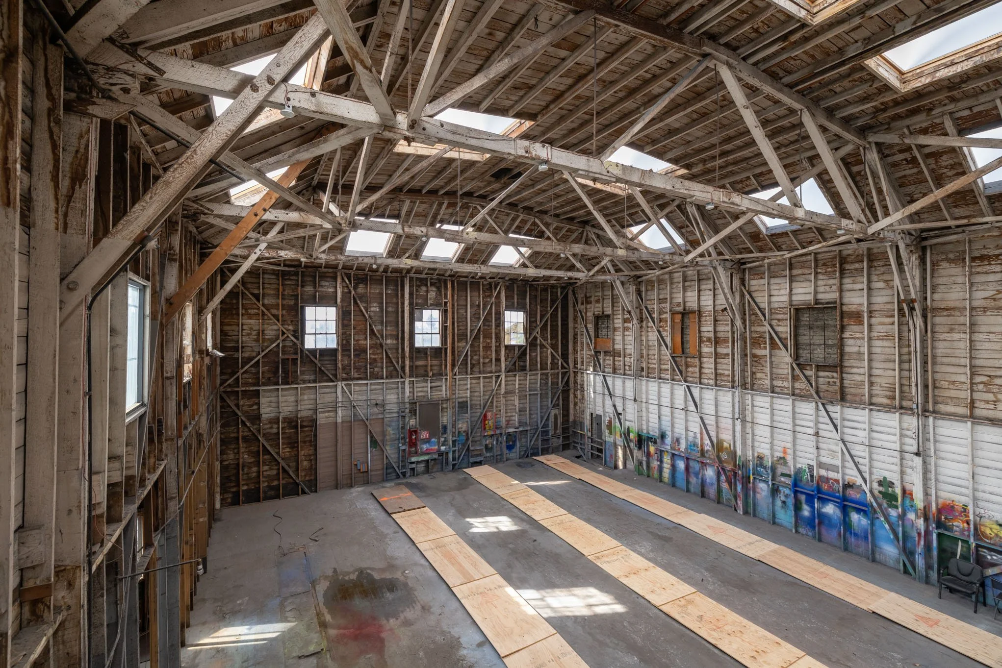 Interior of an unfinished, large, wooden industrial space with exposed beams, partial wall graffiti, and plywood floor sections.