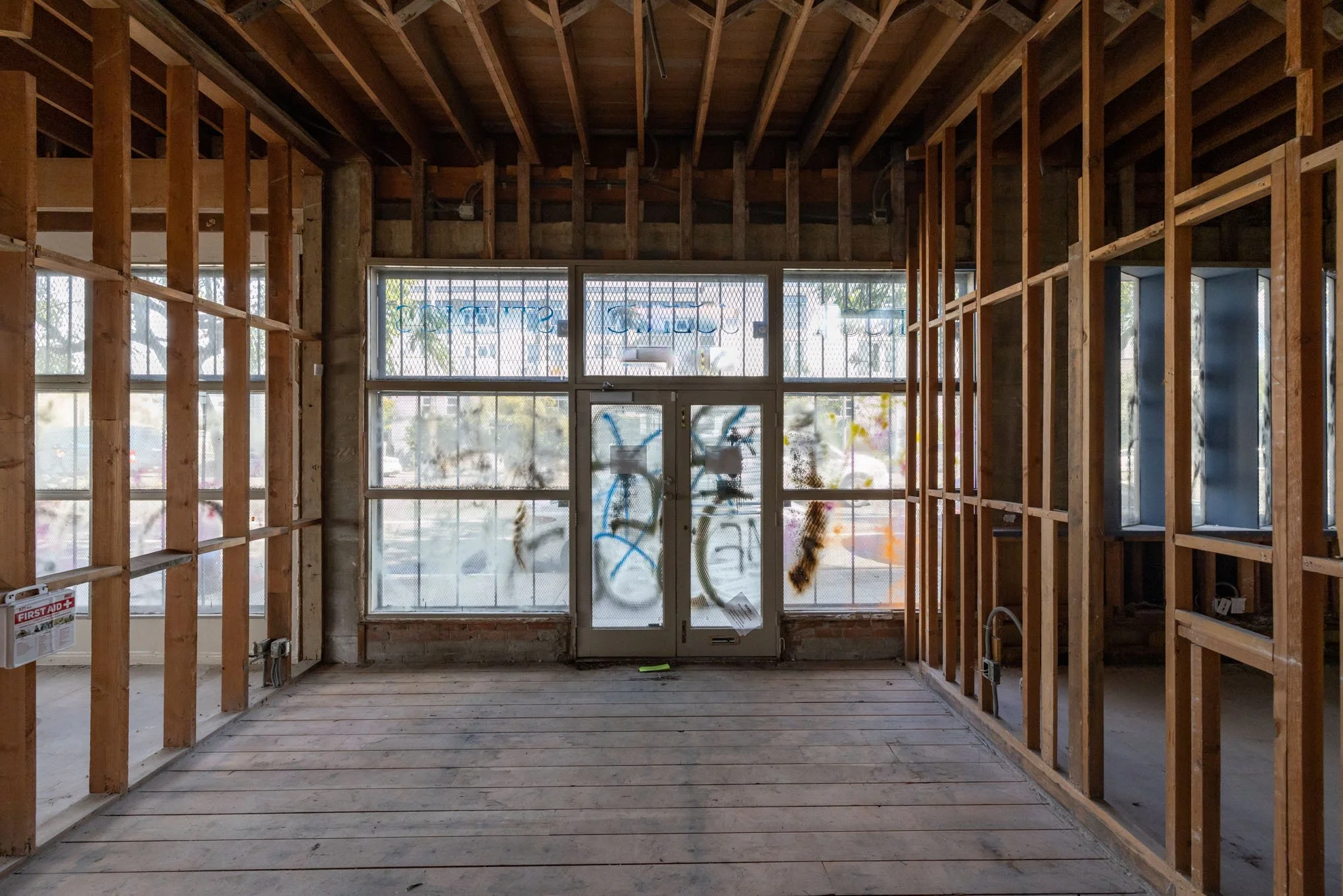 The interior of a building under construction with exposed wooden framing, large front windows with a door, and unfinished flooring.