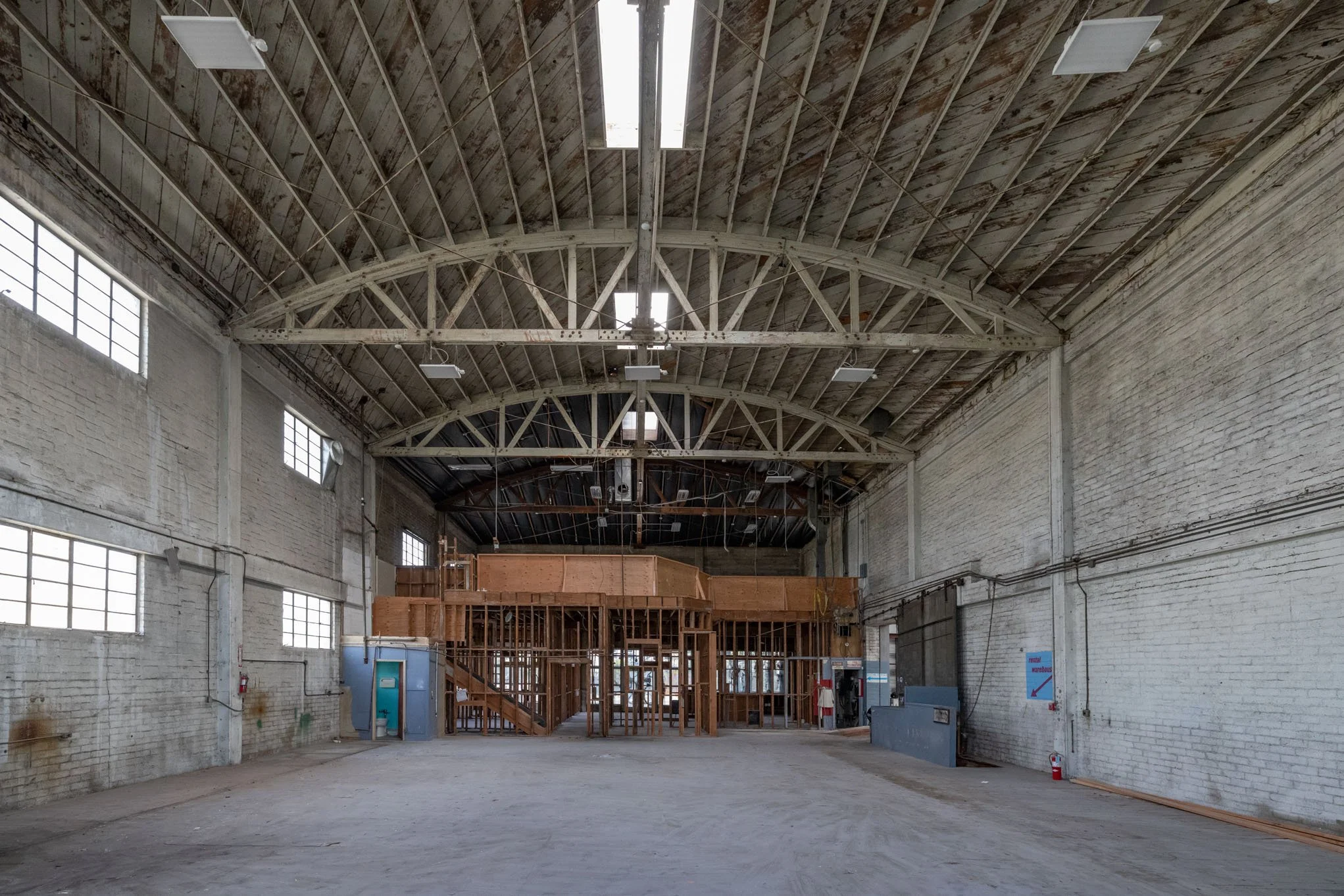 Empty warehouse with high arched ceiling, unfinished wooden structure in the center, and brick walls with windows.