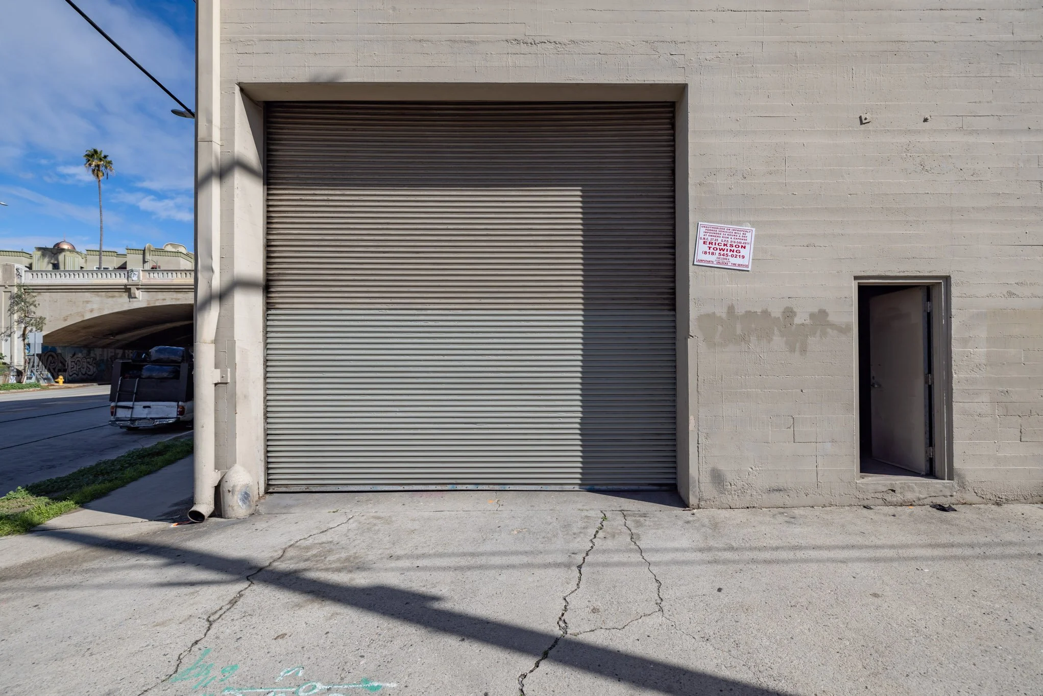 A building with a large closed metal roll-up door, a small open door to the right, a sign above the small door, and a cracked concrete sidewalk in front. A vehicle is partially visible on the street to the left, with a palm tree and clear blue sky in