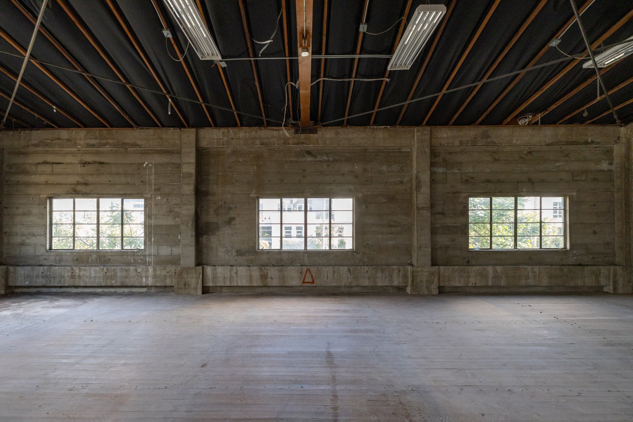 Interior of an unfinished building with concrete walls and floor, three large windows providing daylight, and a ceiling with exposed wooden beams and electrical wiring.