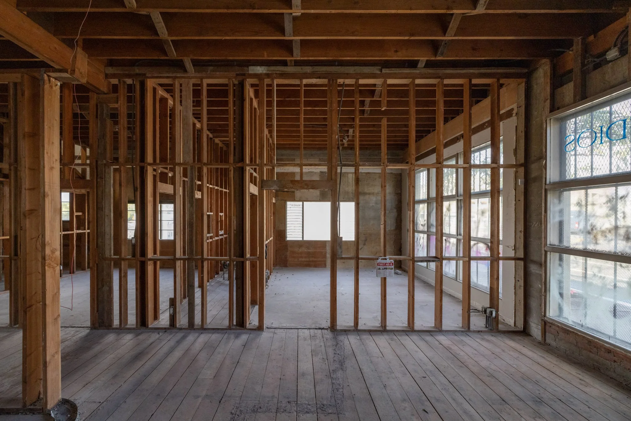 Interior of a building under construction with exposed wooden framing, unfinished walls, and large windows with metal grilles.