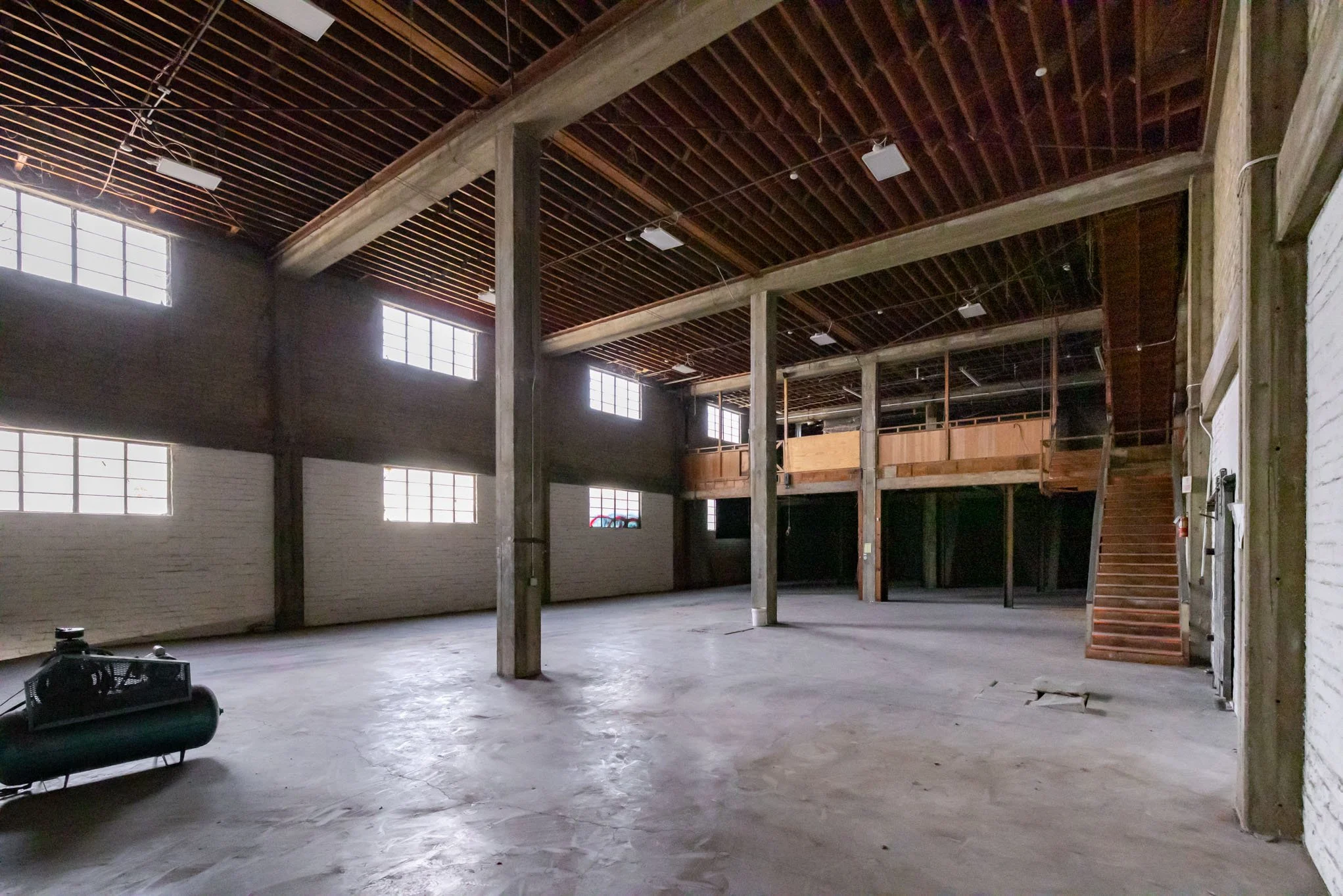 Empty industrial warehouse with concrete floor, brick walls, large windows, wooden beams, and stairs leading to an upper level.